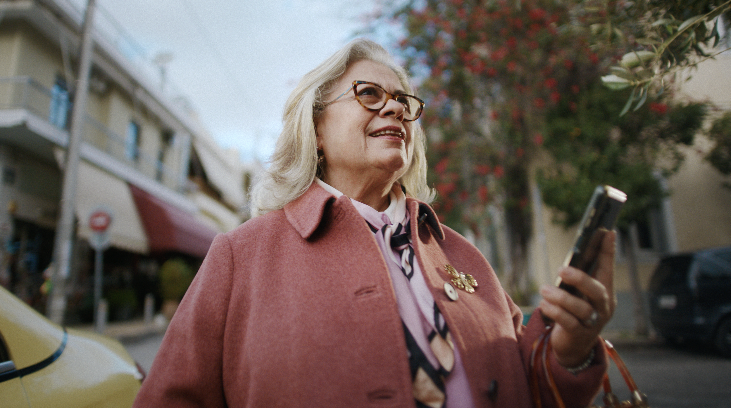 An elderly woman with blonde hair, wearing glasses, a pink coat, and a patterned scarf, holding a phone and smiling while outdoors on a city street with buildings, trees with red leaves, and a yellow taxi in the background.