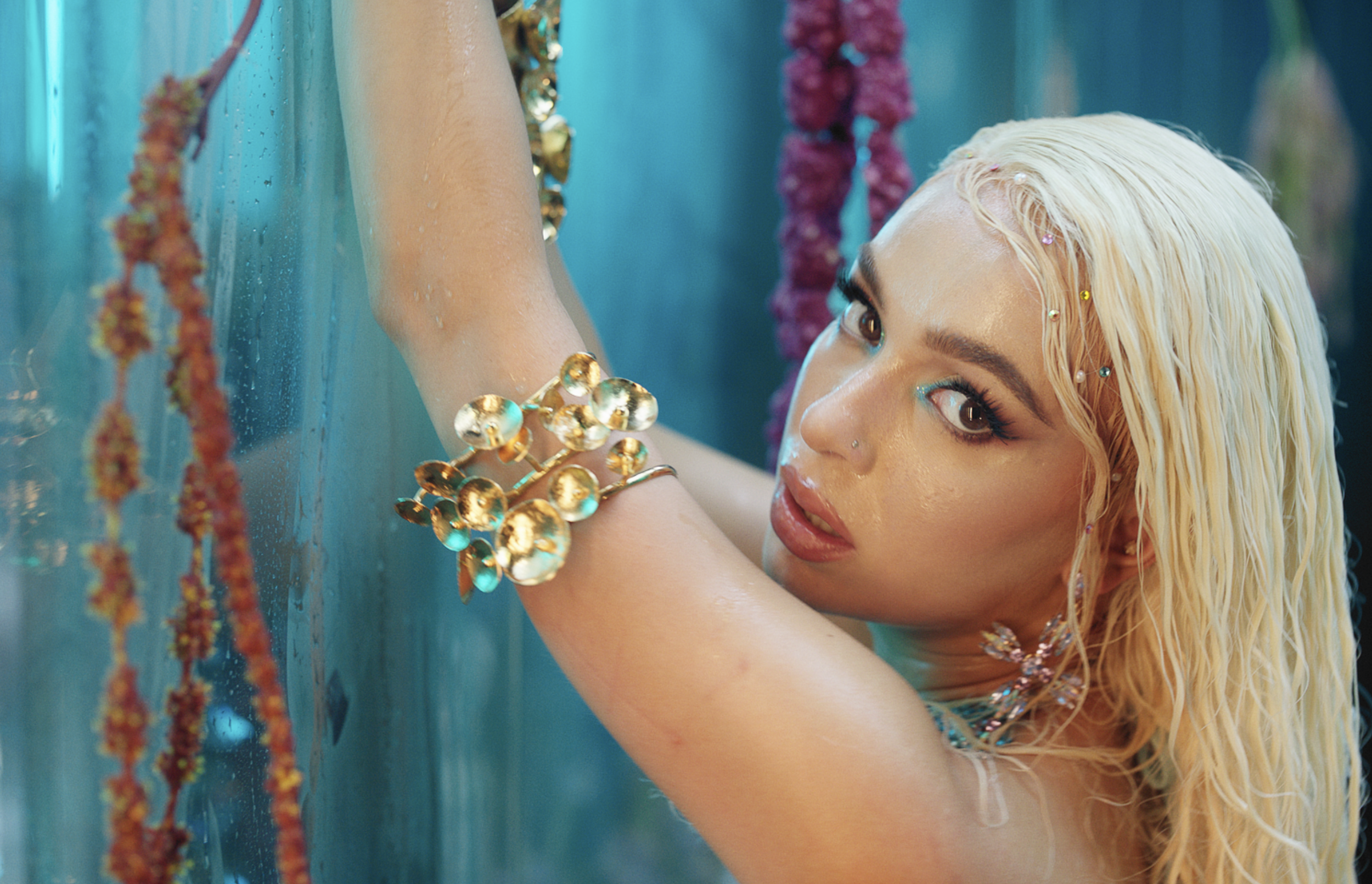 A woman with platinum blonde hair and colorful jewelry resting her head and arm on a waterlogged blue surface, with beads and garlands hanging nearby.