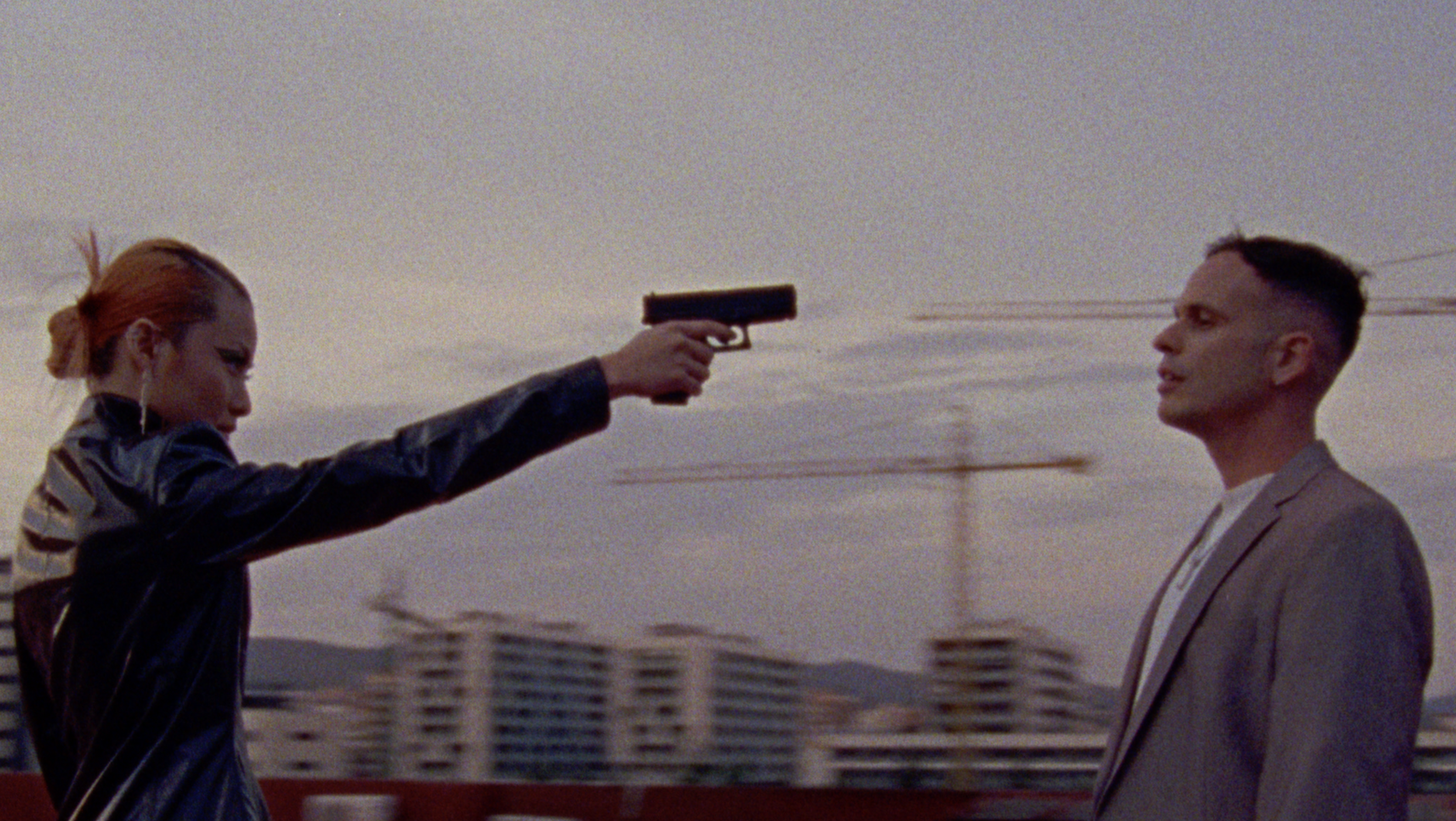 A woman with red hair holding a handgun pointed at a man in a suit, with a cityscape and power lines in the background.