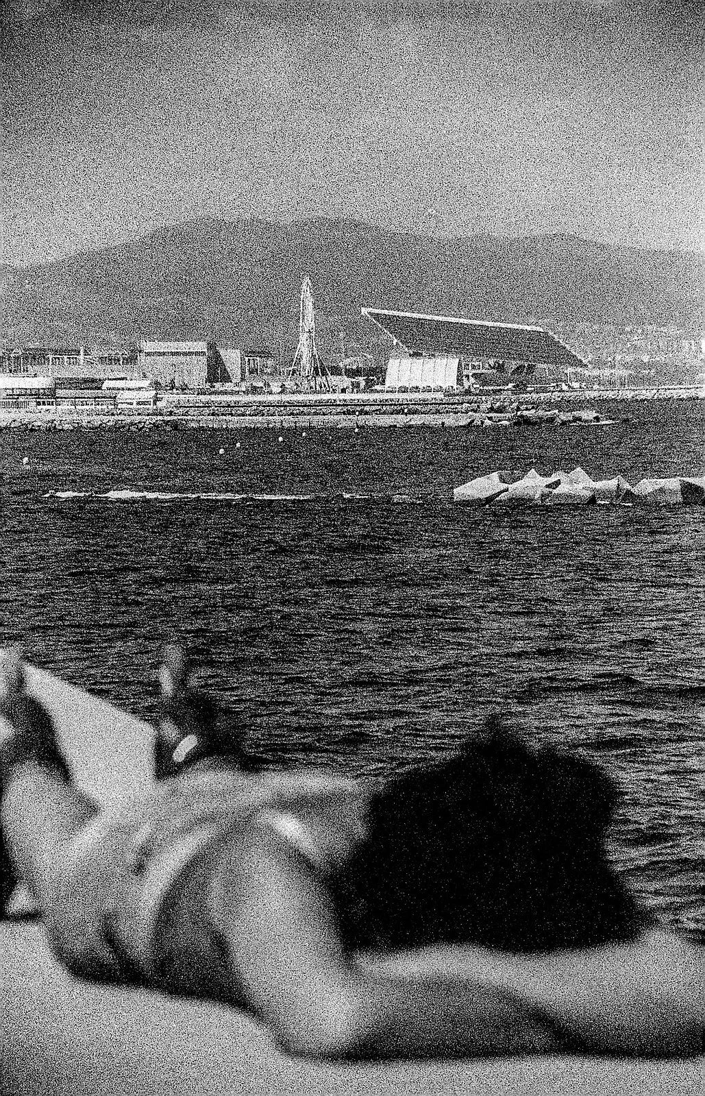 Black and white photo of a waterfront with mountains in the background, a fairground with a Ferris wheel, and a large modern building with an angled roof in the midground. In the foreground, a person is lying down, partially visible and out of focus.