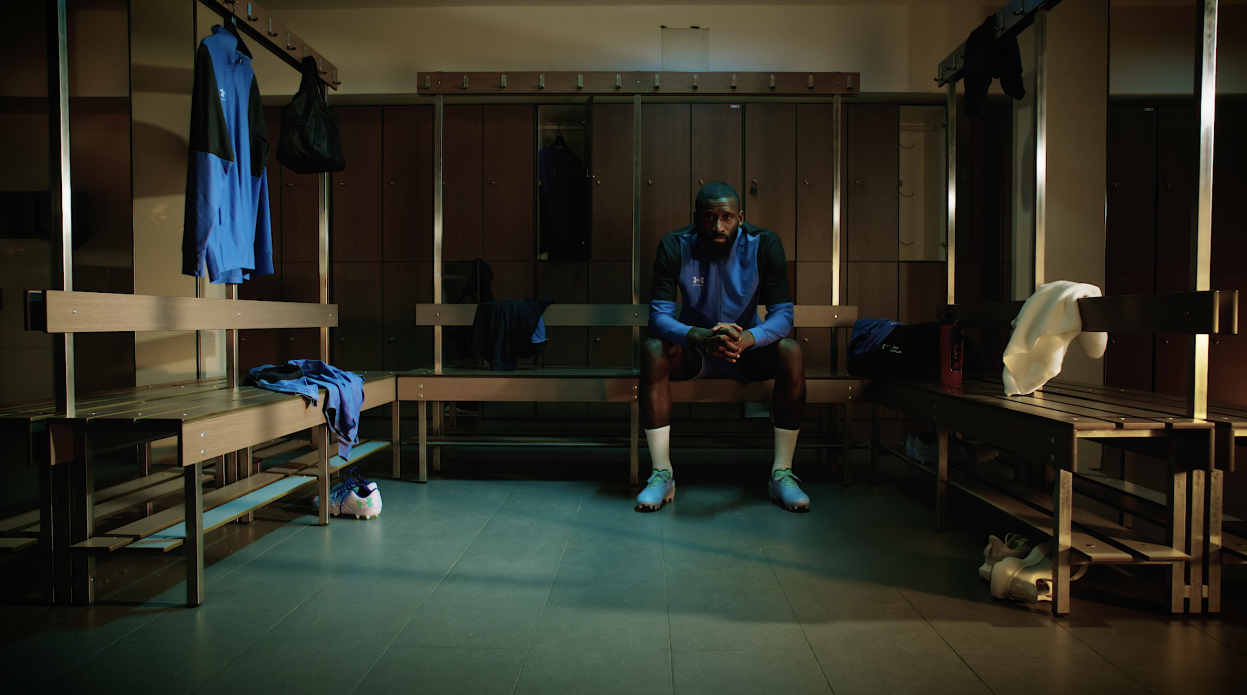 A man sitting on a bench in a locker room, surrounded by lockers and sports gear, wearing athletic clothing and cleats.