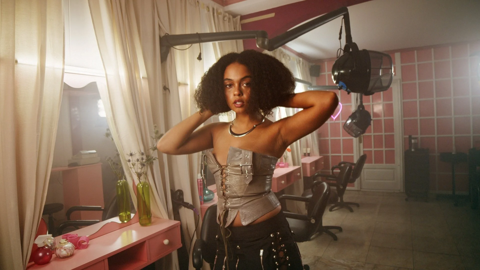 A woman with curly hair posing with her hands behind her head in a hair salon, wearing a strapless leather top and black pants, surrounded by salon furniture and decorations.
