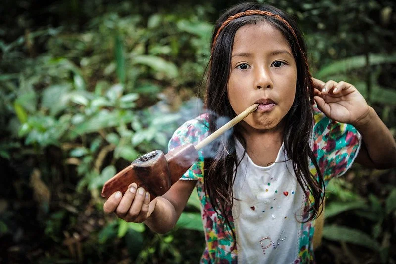 A young girl with long dark hair, wearing a colorful shirt, is smoking a pipe in a lush green forest.