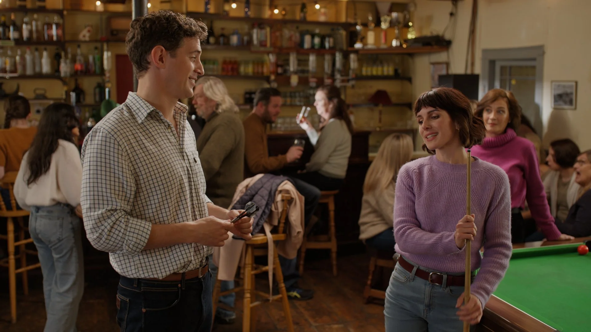 People socializing in a cozy bar or pub, with two women near a pool table, one holding a pool cue and the other holding a pool ball, while others sit and stand around in the background.