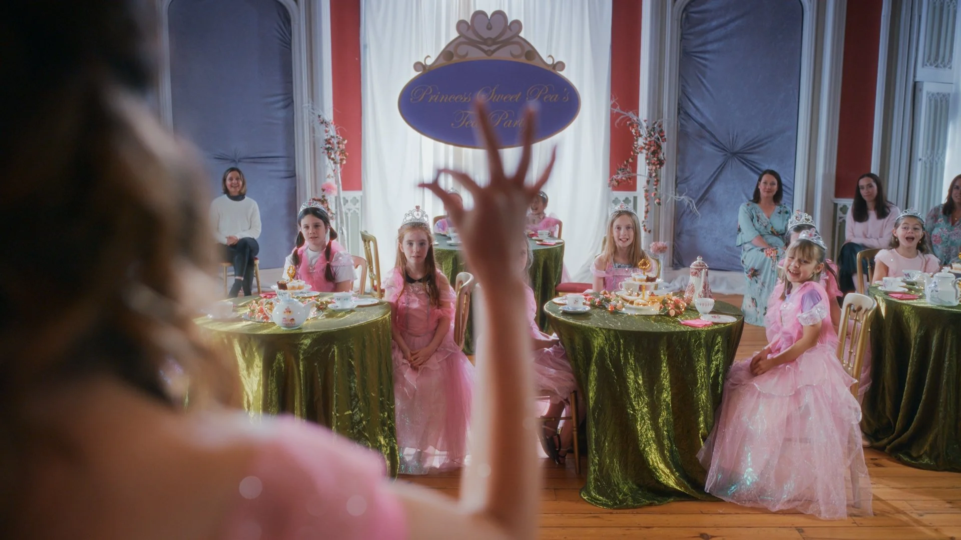 Young girl dressed as a princess at a tea party in a decorated room with princess-themed tables, and children and adults sitting in the background.