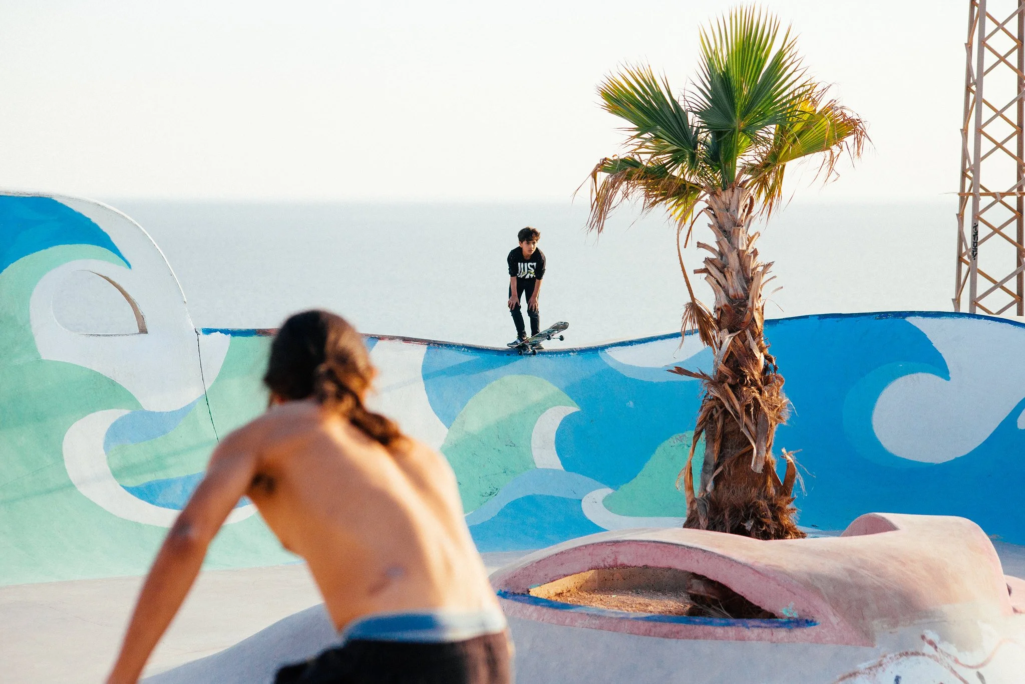 A boy skateboarding on a colorful skatepark with wave murals and a palm tree, with other people in the foreground.