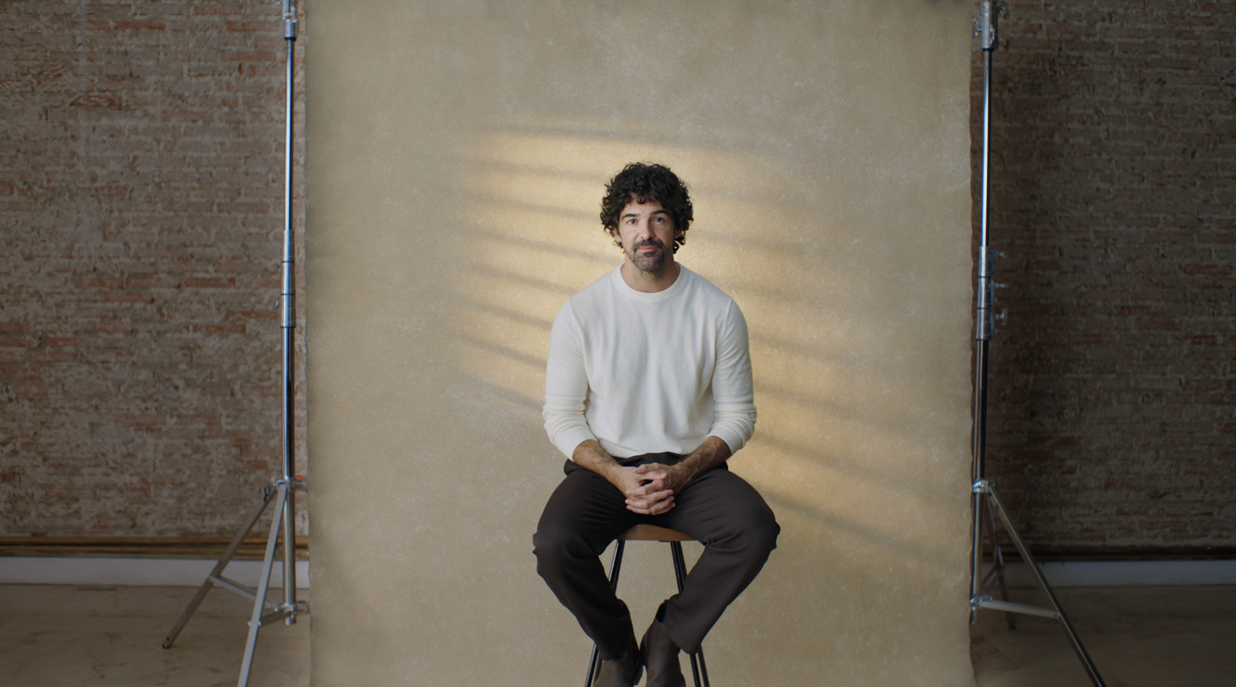 A man with curly dark hair and a beard, wearing a white sweater and dark pants, sitting on a stool in front of a beige backdrop with light patterns on the wall behind him.