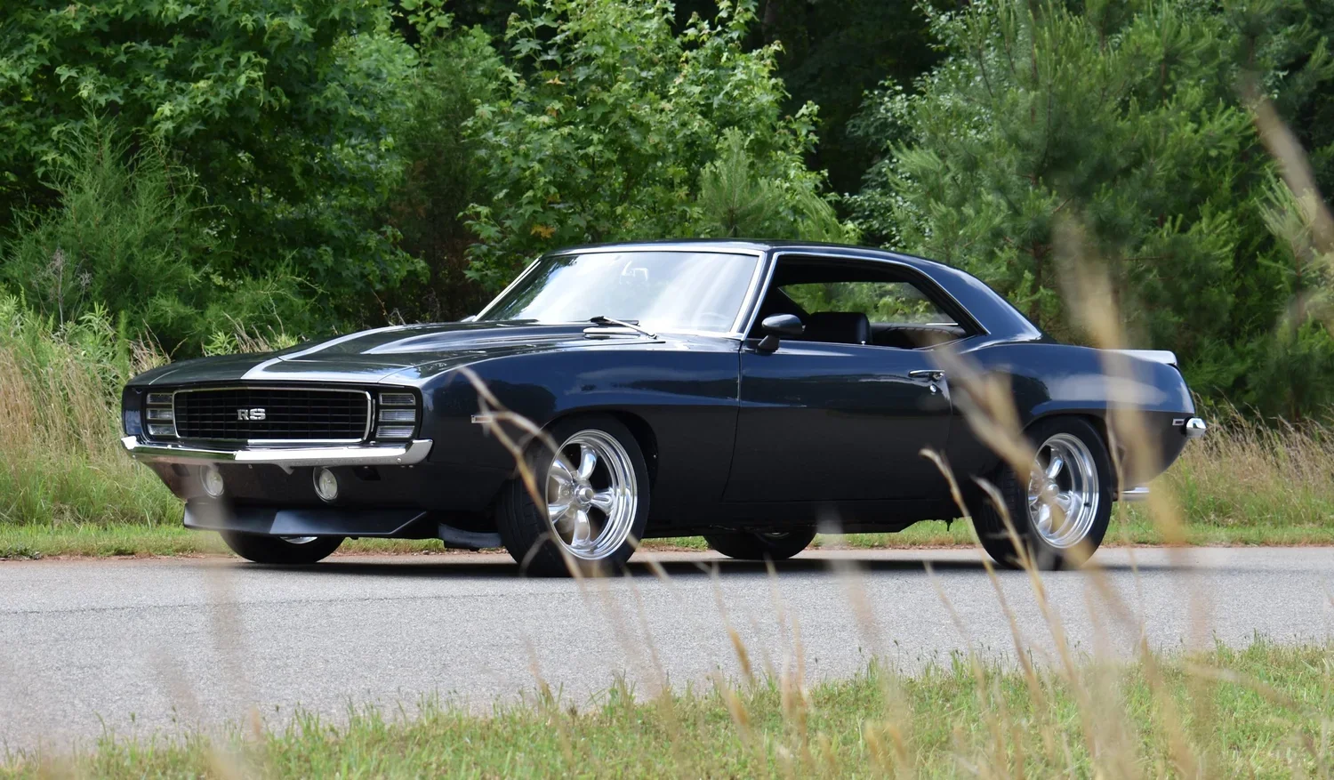 A vintage black Chevrolet Camaro RS car parked on a road with lush green trees and grass in the background.