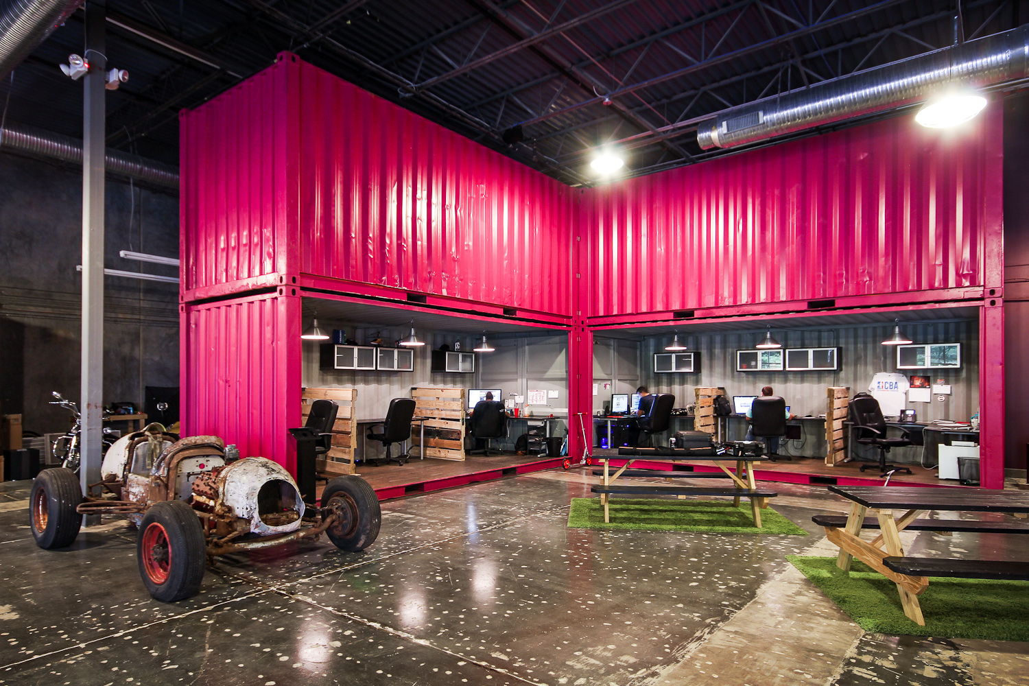 Industrial office space inside a repurposed shipping container with four people working at computers. The space has pink walls and outdoor furniture on the floor, with an old, rusted car chassis in the foreground.