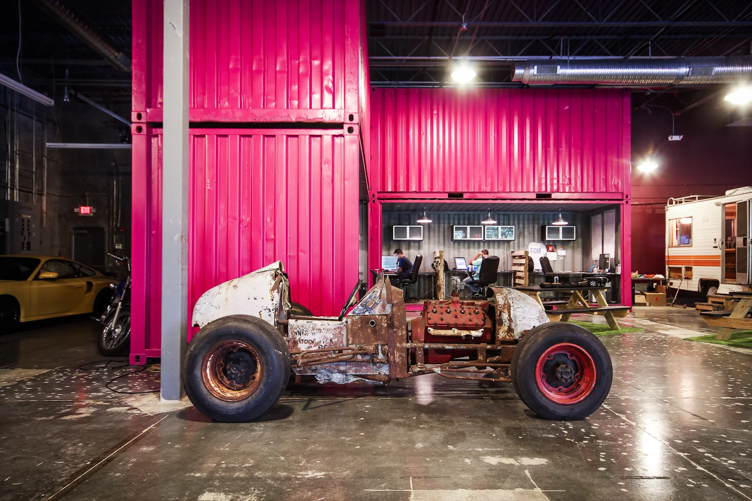 Old, rusted car chassis in front of a bright pink wall inside an industrial-style space, with people working at desks in a small office area and a yellow sports car and motorcycle to the side.