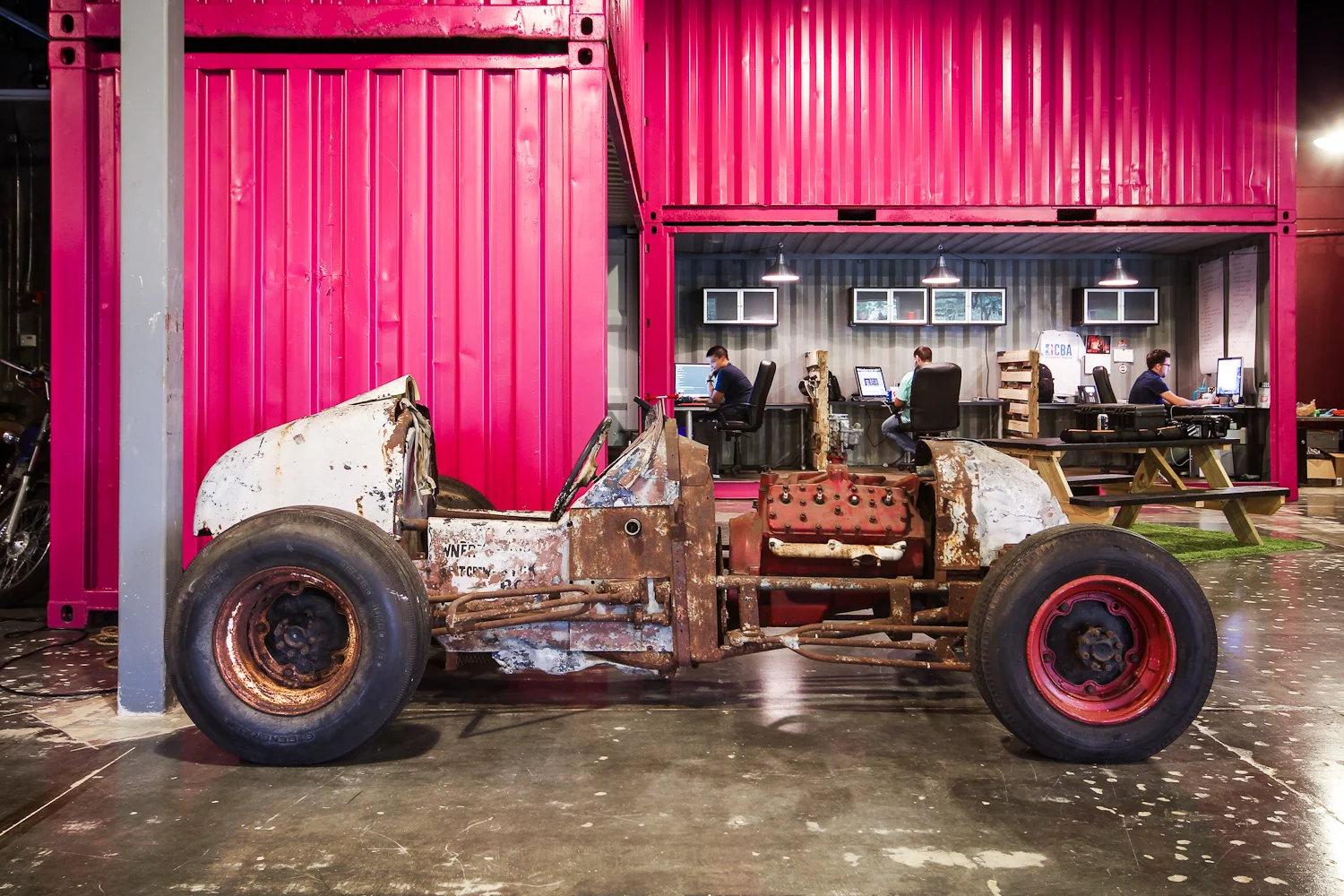 A rusted vintage race car body with missing parts and peeling paint, positioned indoors in front of a pink shipping container with an office area behind it where three people are working at computers.