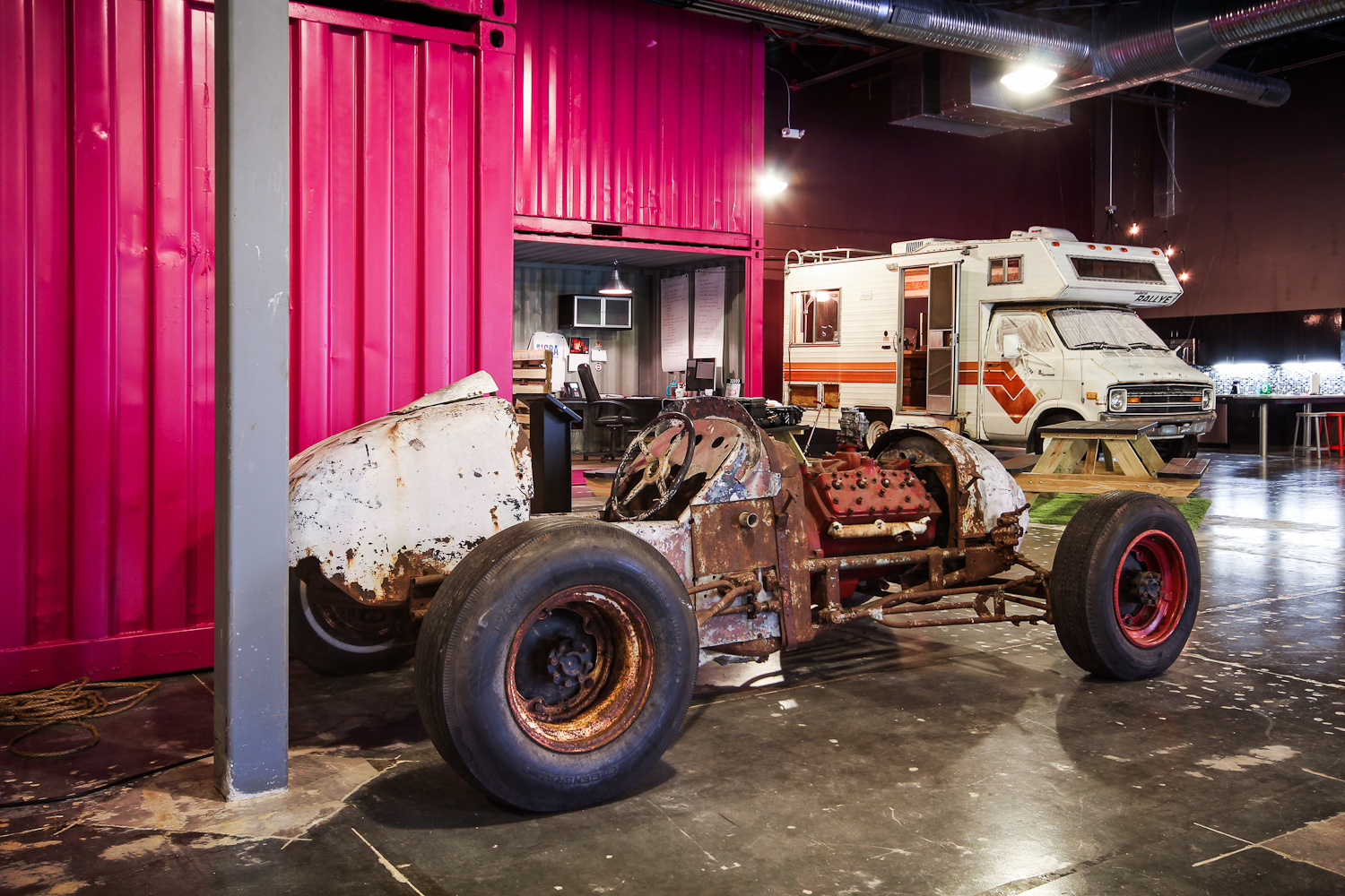 Inside a colorful vehicle museum displaying a rusty race car chassis, a vintage RV, and a partially visible pink container with a small office in the background.