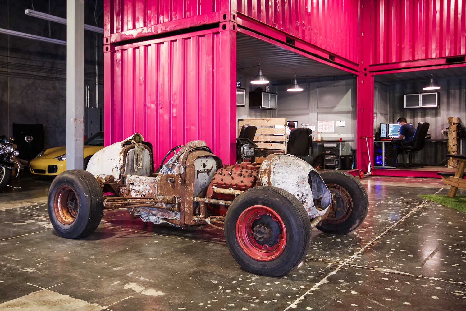 An unfinished, rusty vintage race car body with no paint, mounted on a frame with large tires, inside a modern industrial garage. Behind it, there are garages with a yellow sports car, and workstations with two people working on computers.