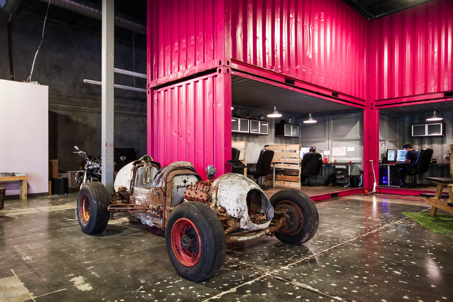 Rusty car frame inside an industrial-style office space with bright pink storage containers and a few employees working at computers.