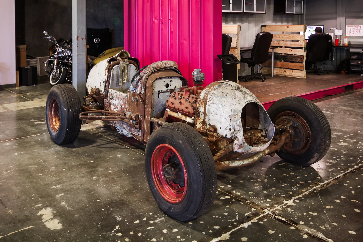 A rusty, incomplete vintage car frame on a concrete floor, with a motorcycle and an office space with desks and chairs in the background.