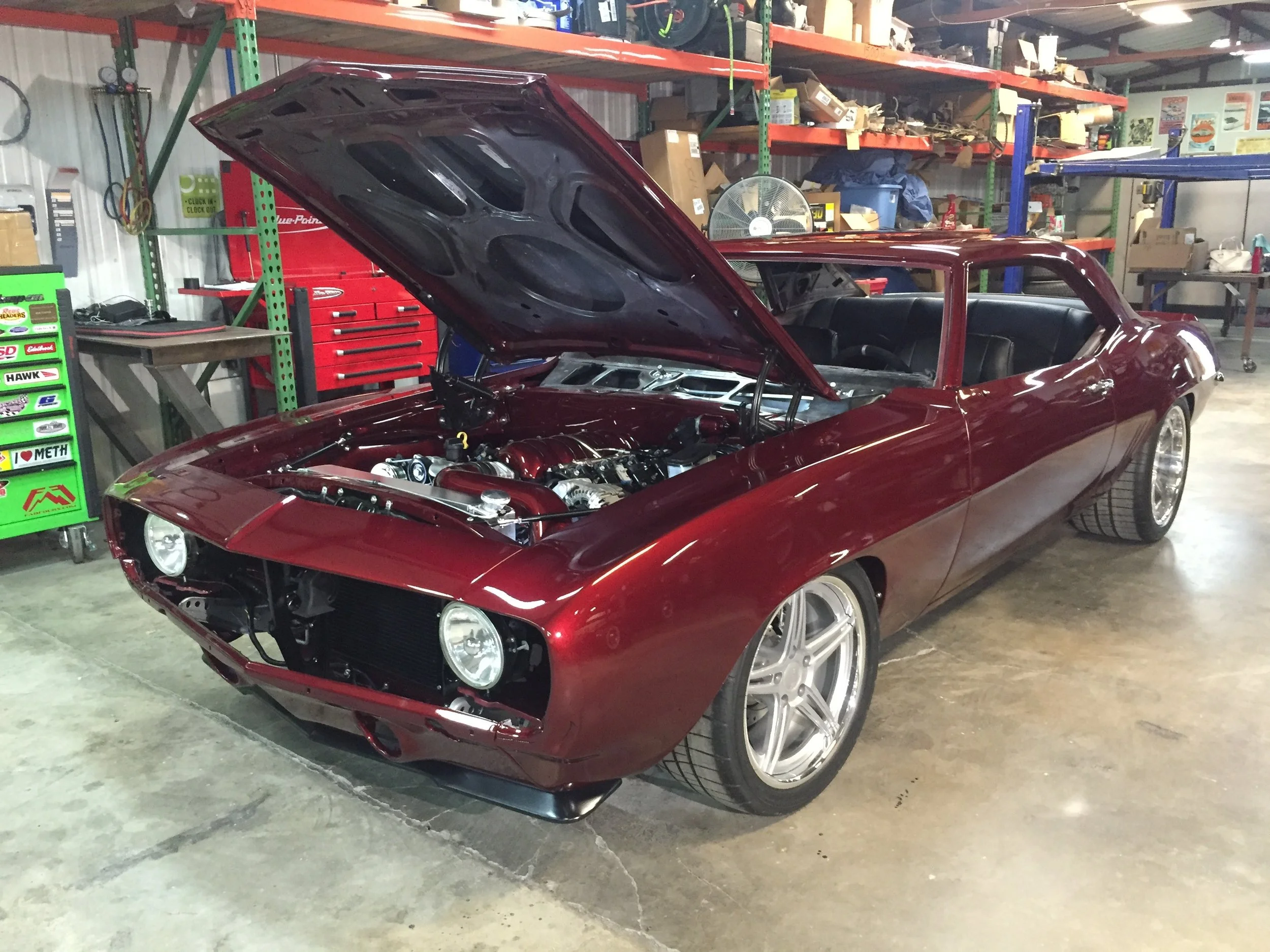 A red classic car with its hood open, inside a garage or workshop with shelves containing various tools, boxes, and equipment.
