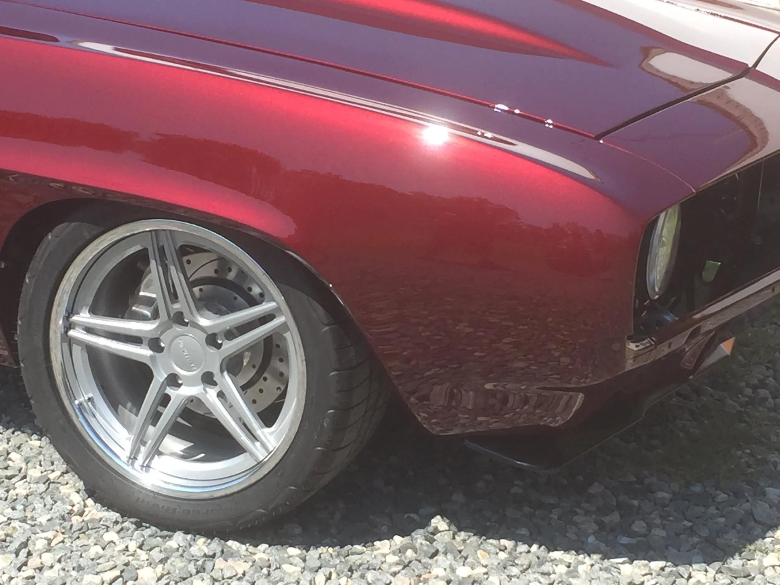 A close-up of a red sports car, showing the front left wheel and part of the wheel well, with reflections on the shiny car surface and a gravel ground underneath.