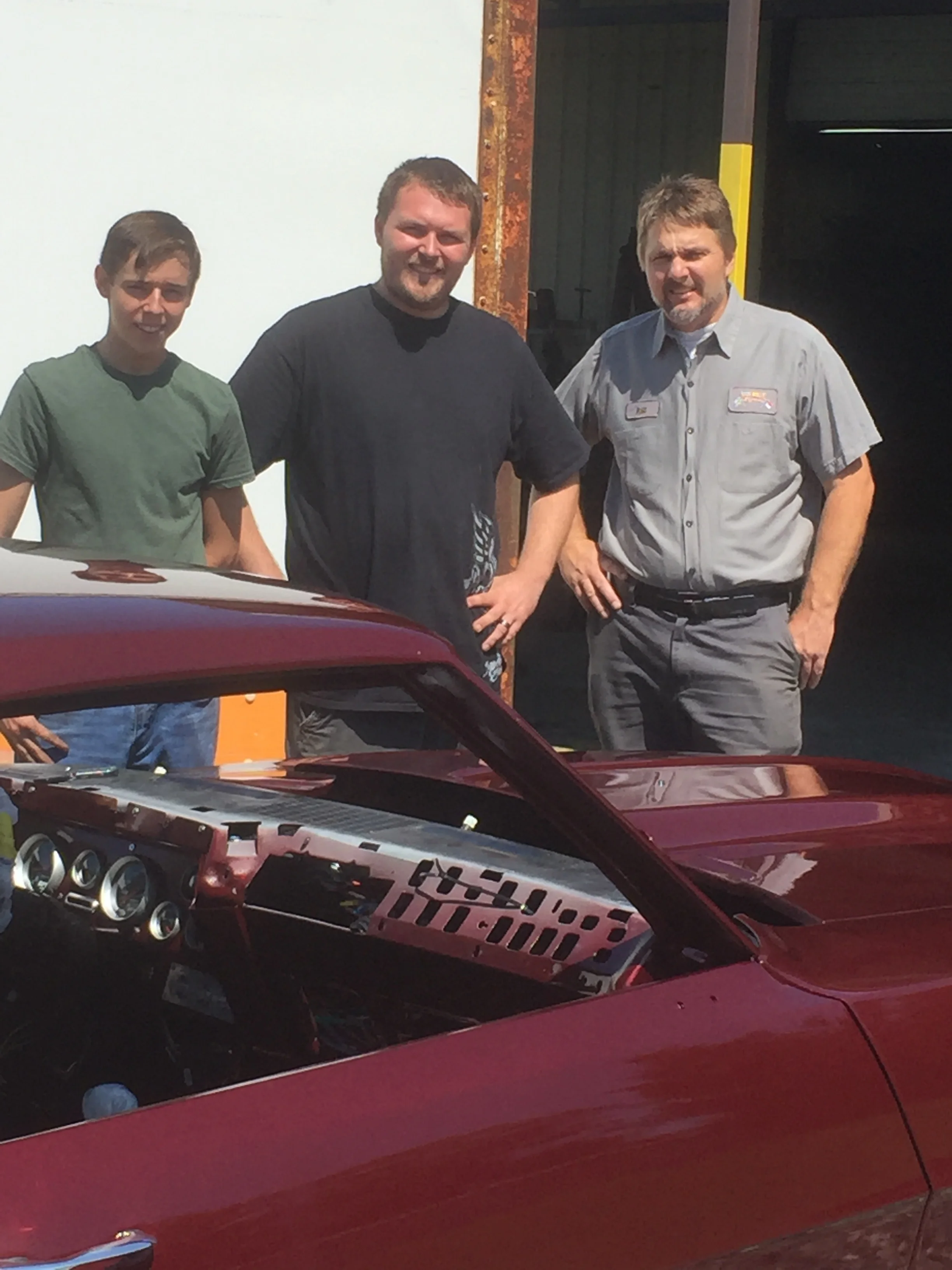 Three men standing outdoors in front of a white wall and a black structure, with a red car in the foreground.