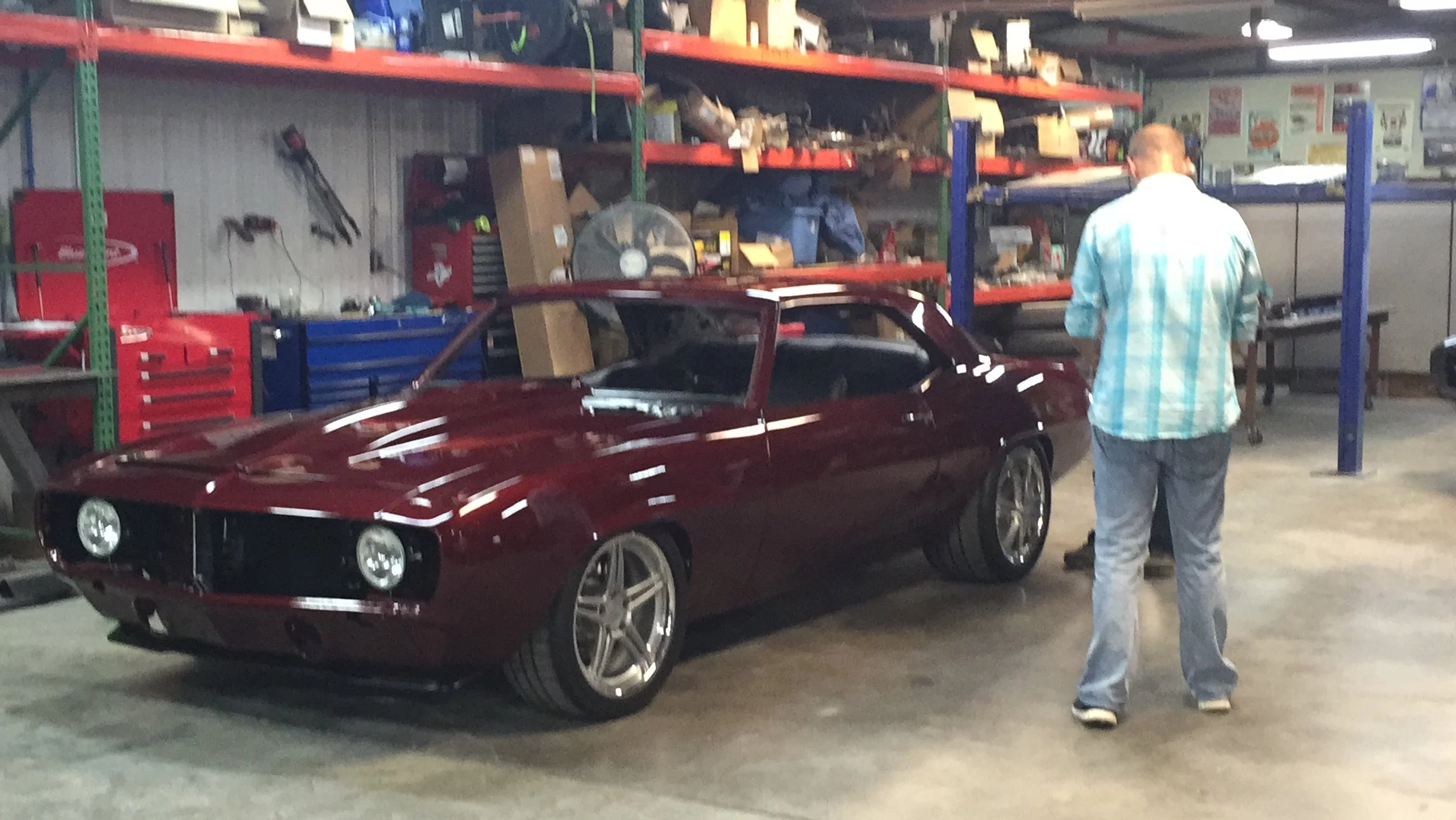 A person with a plaid shirt and jeans stands near a shiny maroon sports car inside a garage or workshop with shelves filled with tools and parts.