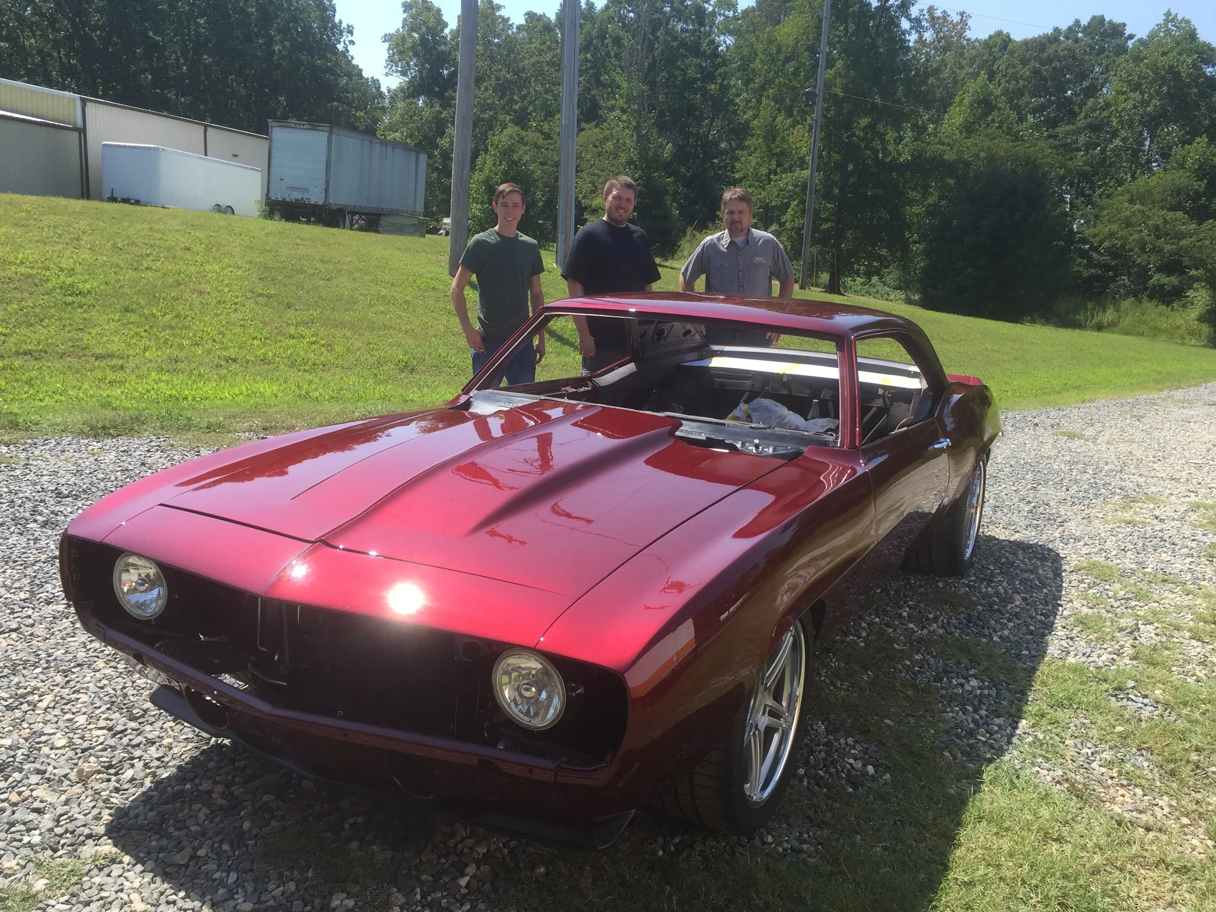 Three men standing behind a red muscle car with an open hood, outdoors on a sunny day, with trailer trucks and green trees in the background.
