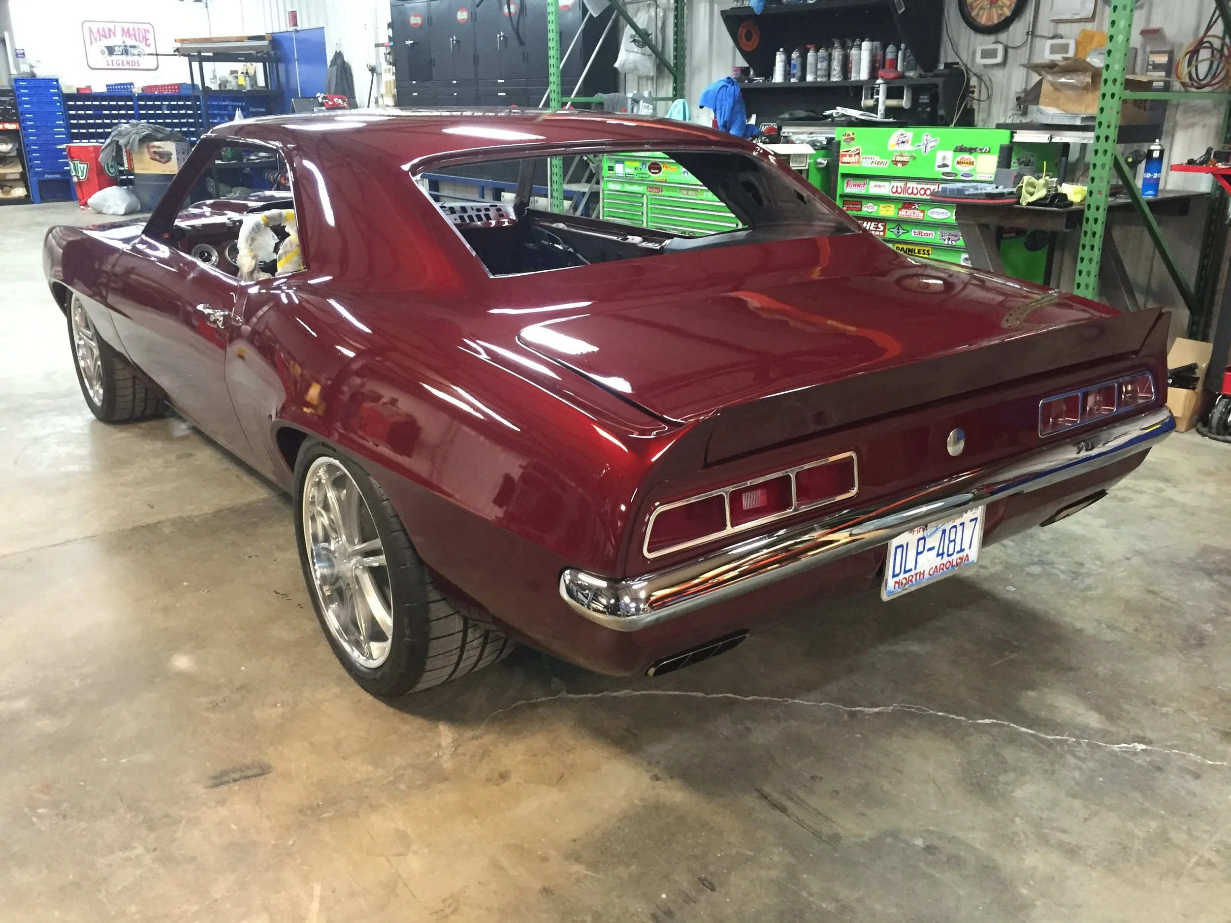 A vintage red muscle car inside a workshop with various tools, shelves, and equipment in the background.
