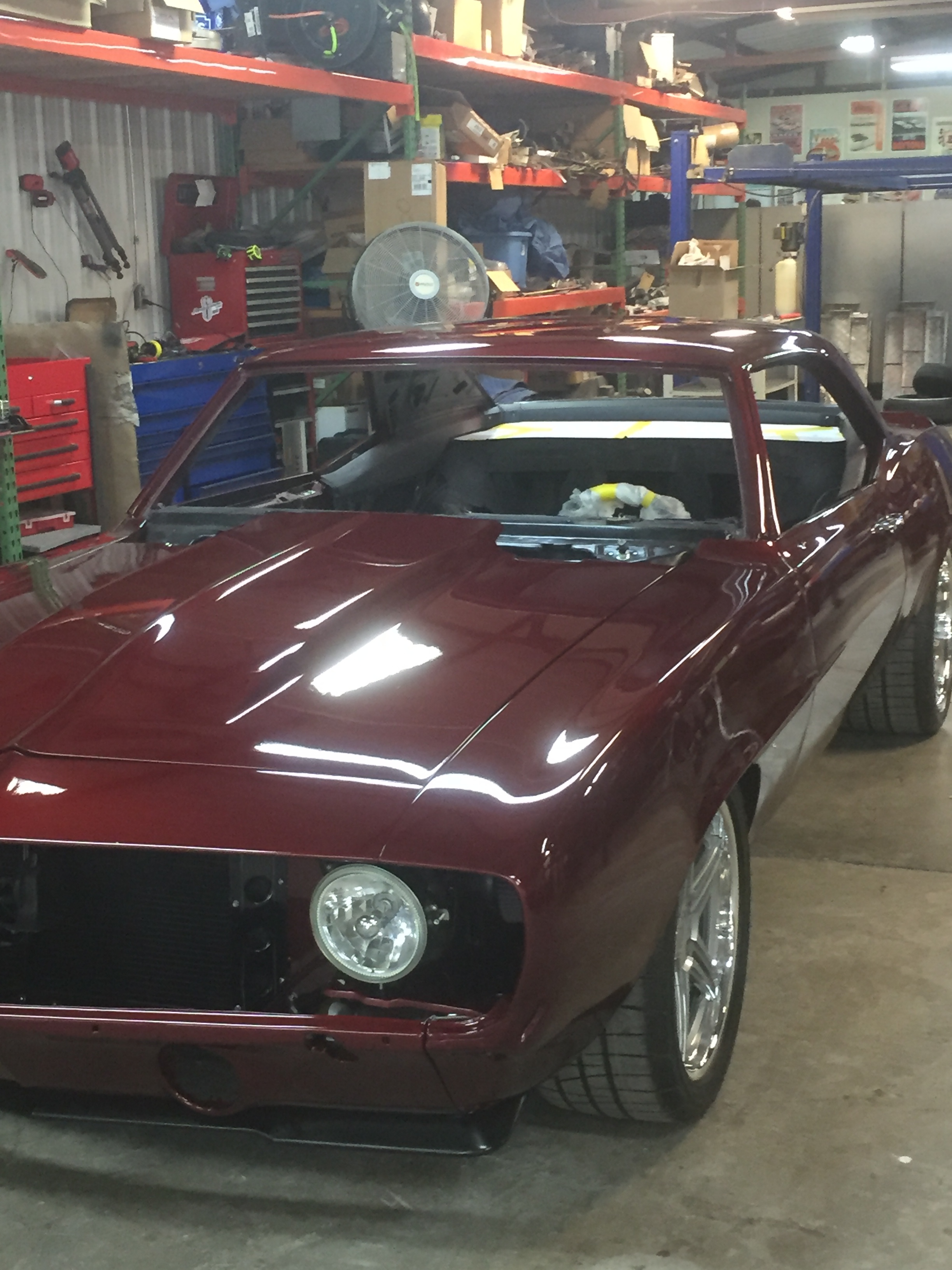 A red vintage sports car is parked inside a cluttered workshop or garage, with various tools and storage shelves visible in the background.