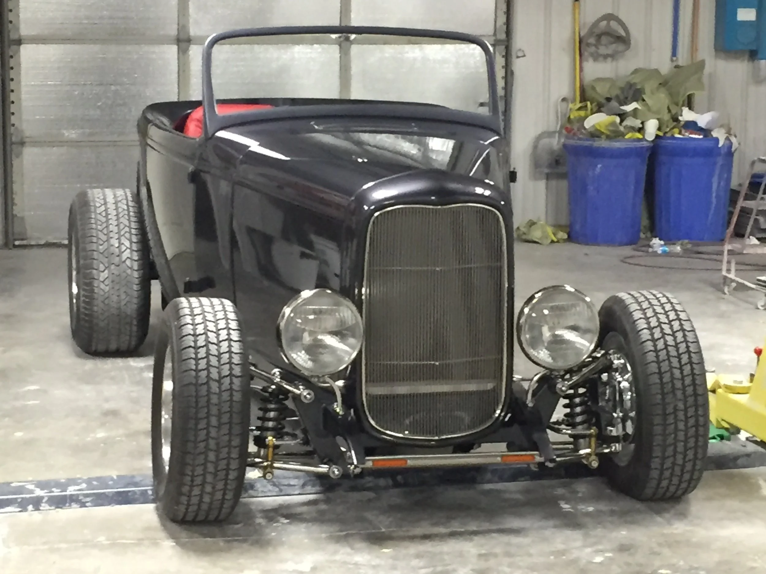 An unfinished black vintage car with no windshield and missing body panels, placed inside a garage with trash bins and tools in the background.