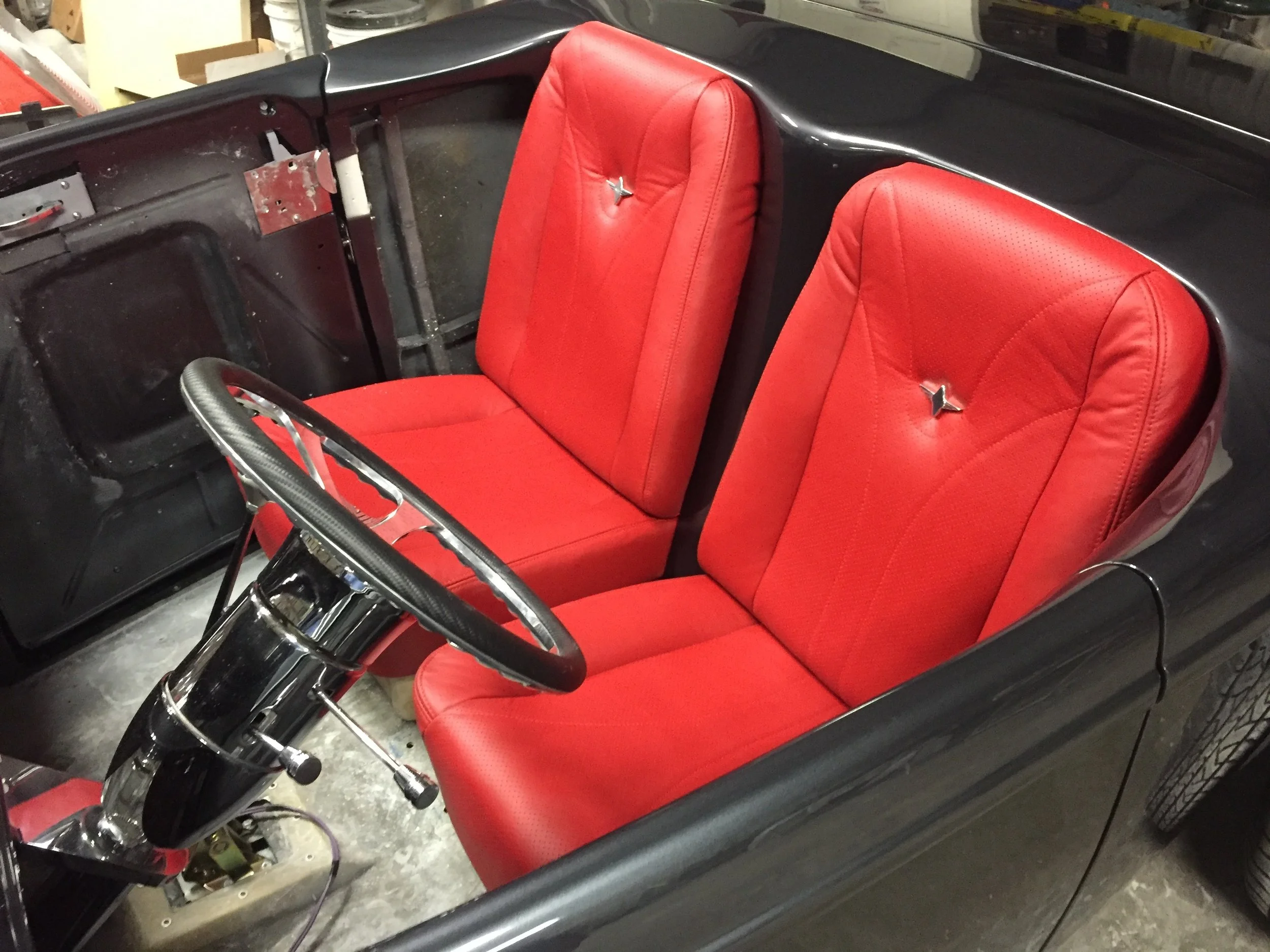 The image shows a vintage golf cart with two red cushioned seats, a black body, and a chrome steering wheel, situated in a garage or workshop.
