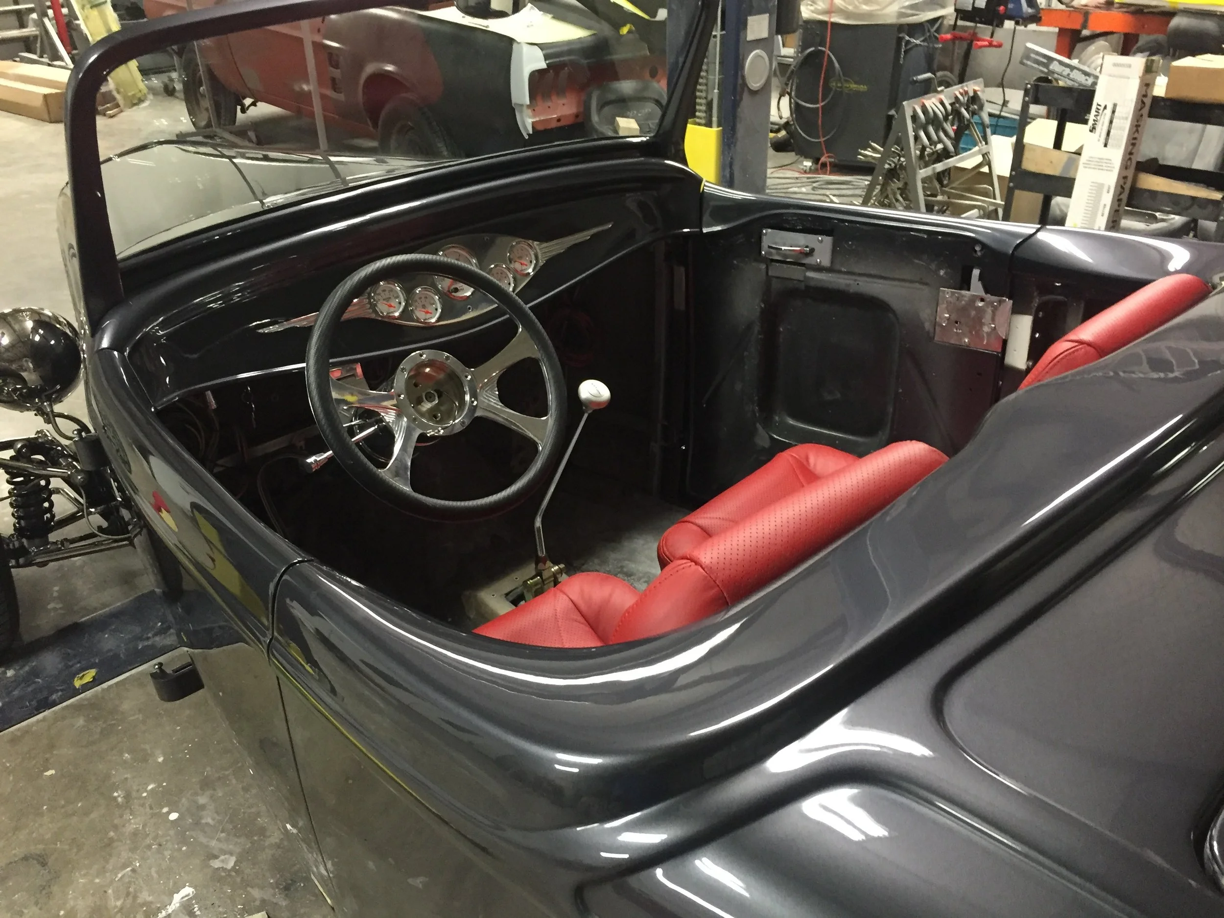 Interior of a vintage race car with red seats, black dashboard with multiple gauges, and gear shift, in a workshop with tools and car parts in the background.