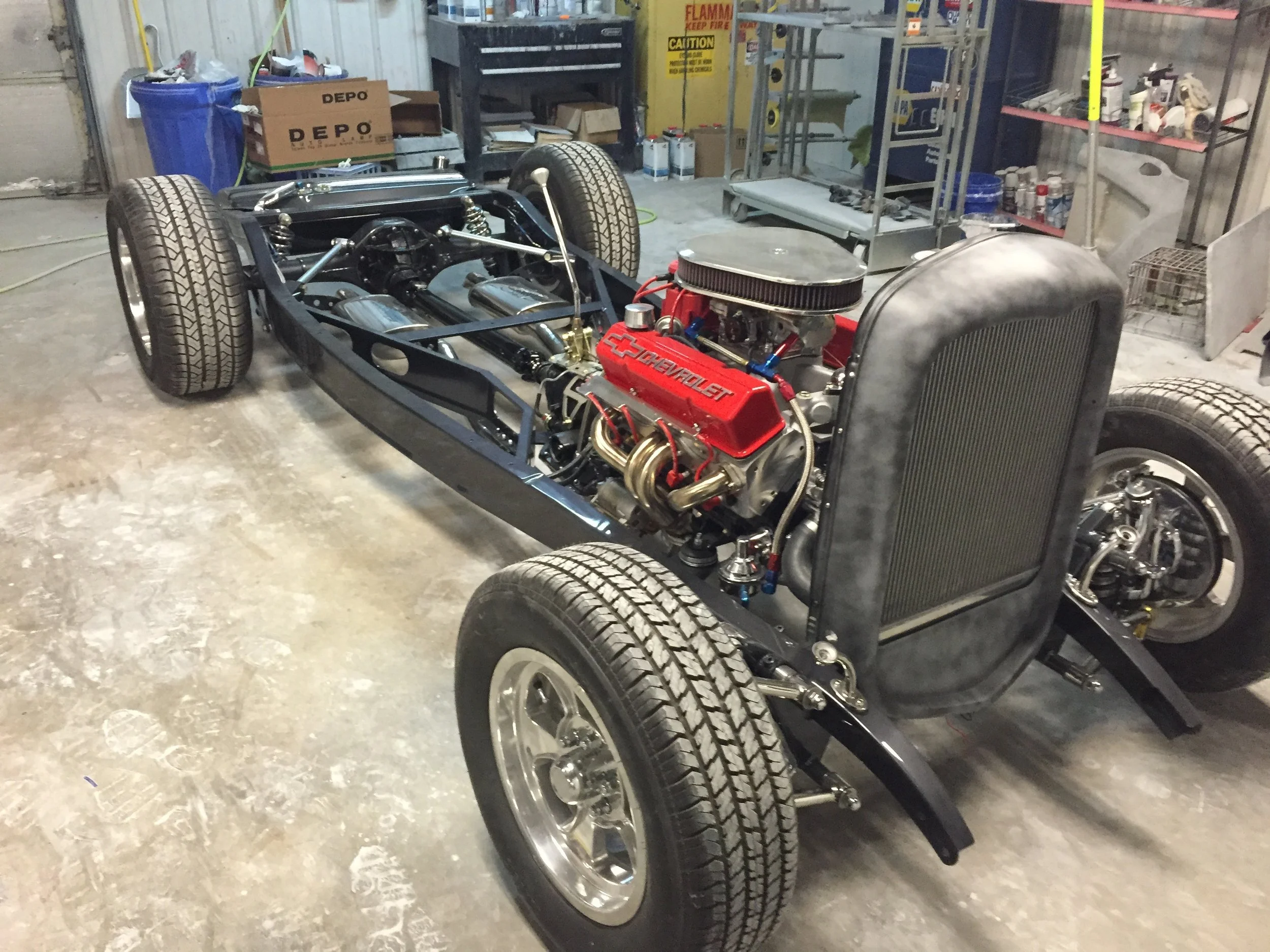 A partially assembled vintage race car with exposed engine and black chassis inside a workshop. The engine has a red Chevrolet logo. The workshop has shelves with tools, supplies, and cardboard boxes, and the floor is concrete.