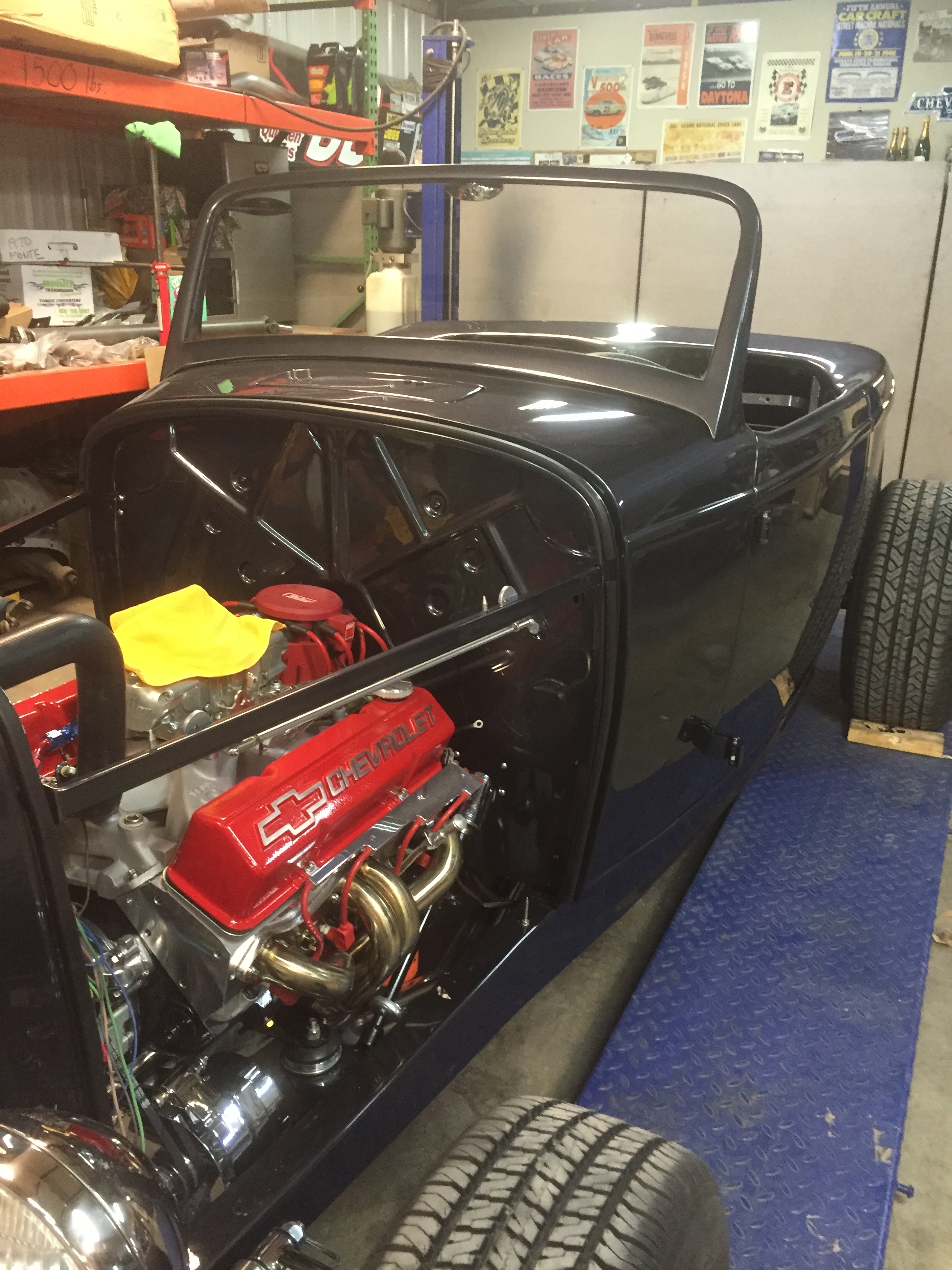 A black vintage car body with a red Chevrolet engine and shiny chrome parts, in a garage with tires and automotive posters on the walls.