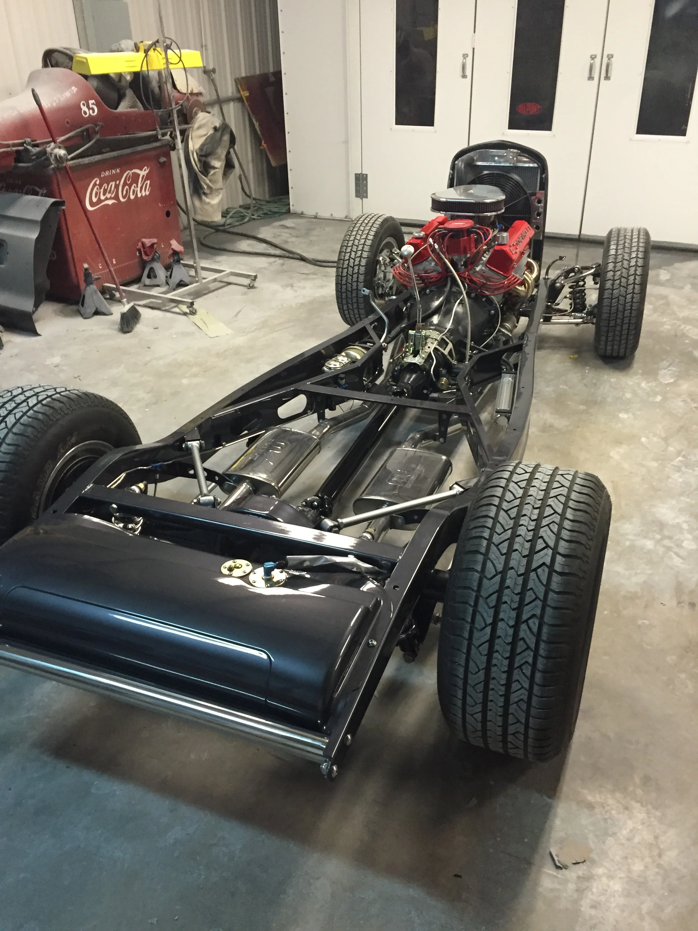 A partially assembled vintage race car with exposed engine and black body, inside a garage with a Coca-Cola tool cart and cleaning supplies nearby.