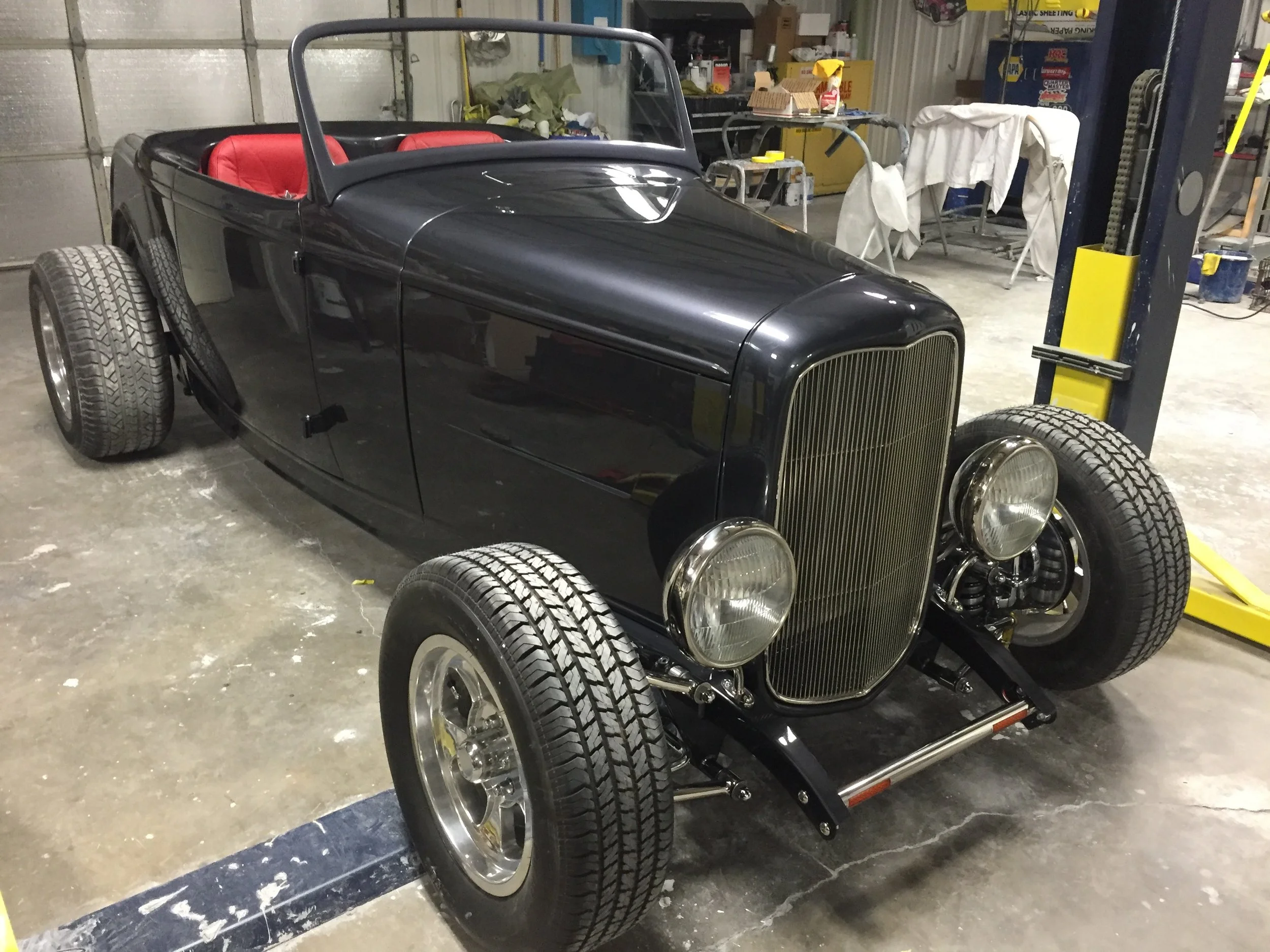 A vintage black roadster car with red interior seats inside a garage or workshop, surrounded by tools and equipment.