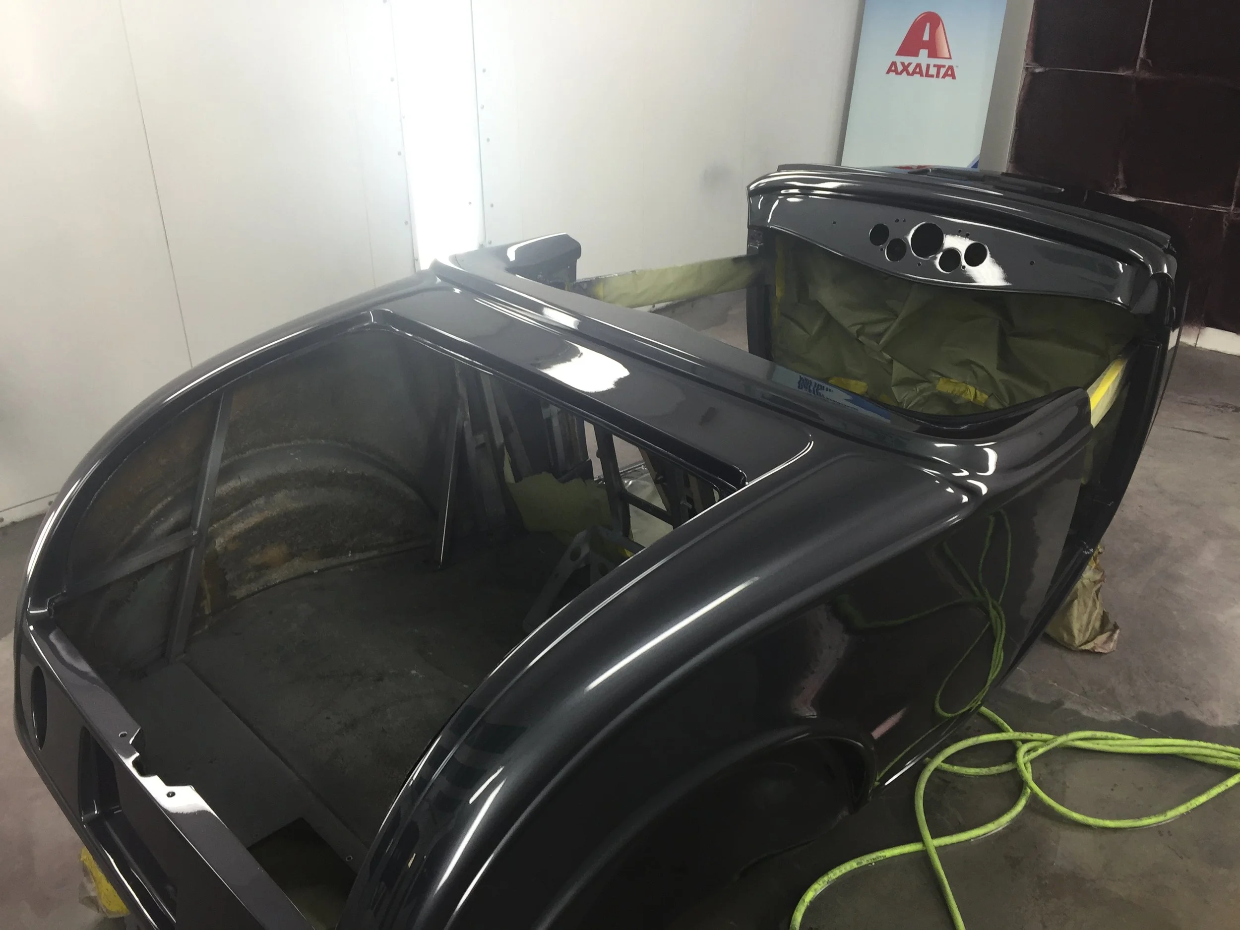 A gray car in a paint booth with its back hatch open, revealing the interior and some painting supplies inside, with a yellow air hose on the floor.
