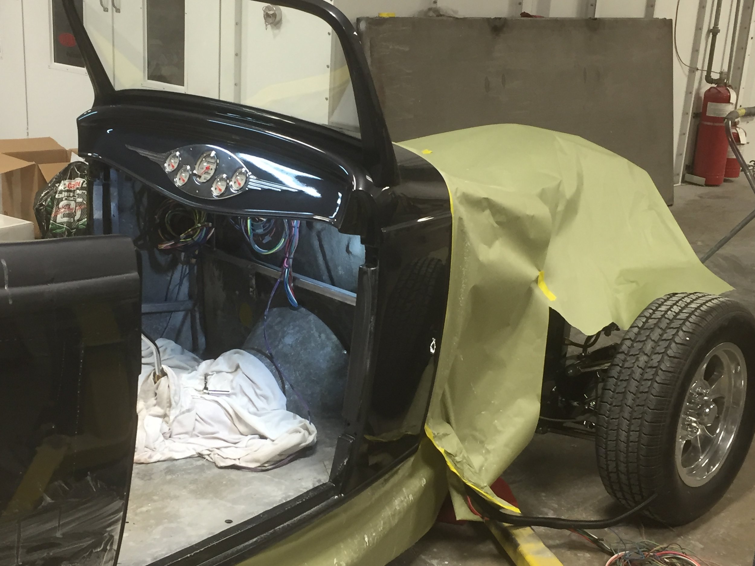 A partially assembled classic black car with a yellow covering over part of the body in a garage or workshop, with tools and supplies around.