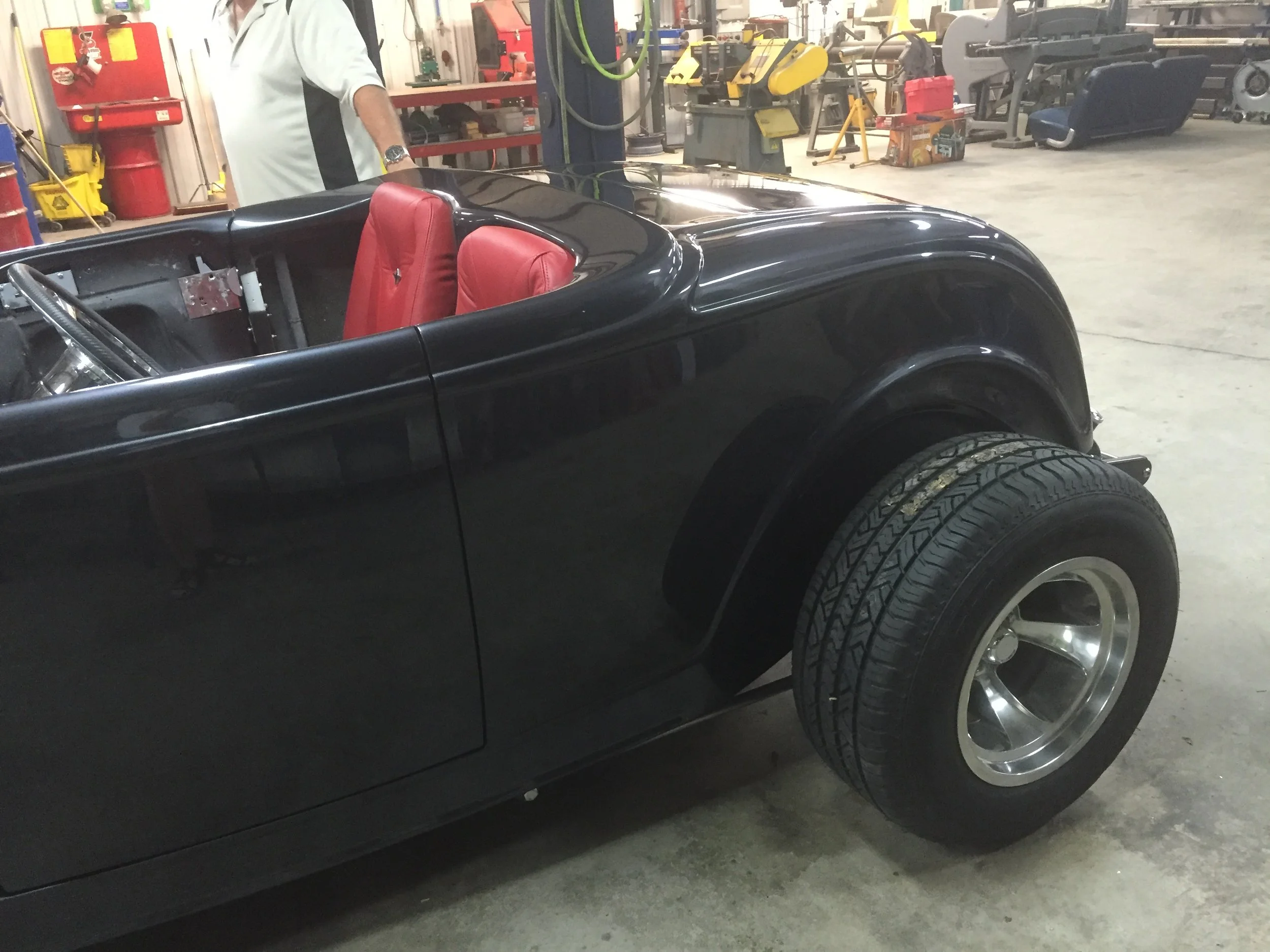 A black vintage convertible car with red leather seats inside a garage workshop, with tools and equipment in the background.