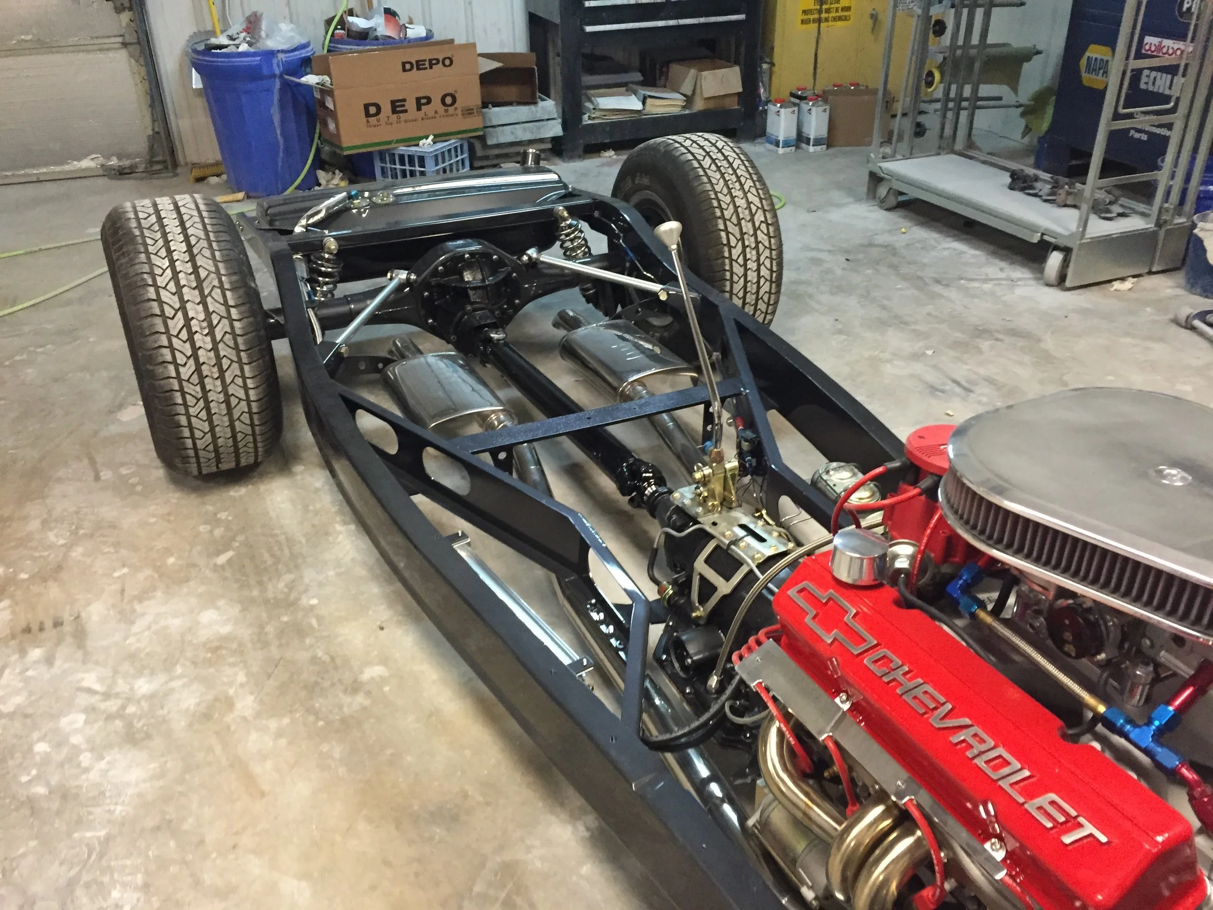 The image shows a partly assembled race car chassis in a garage. The car has large tires, a red engine, and visible suspension components.