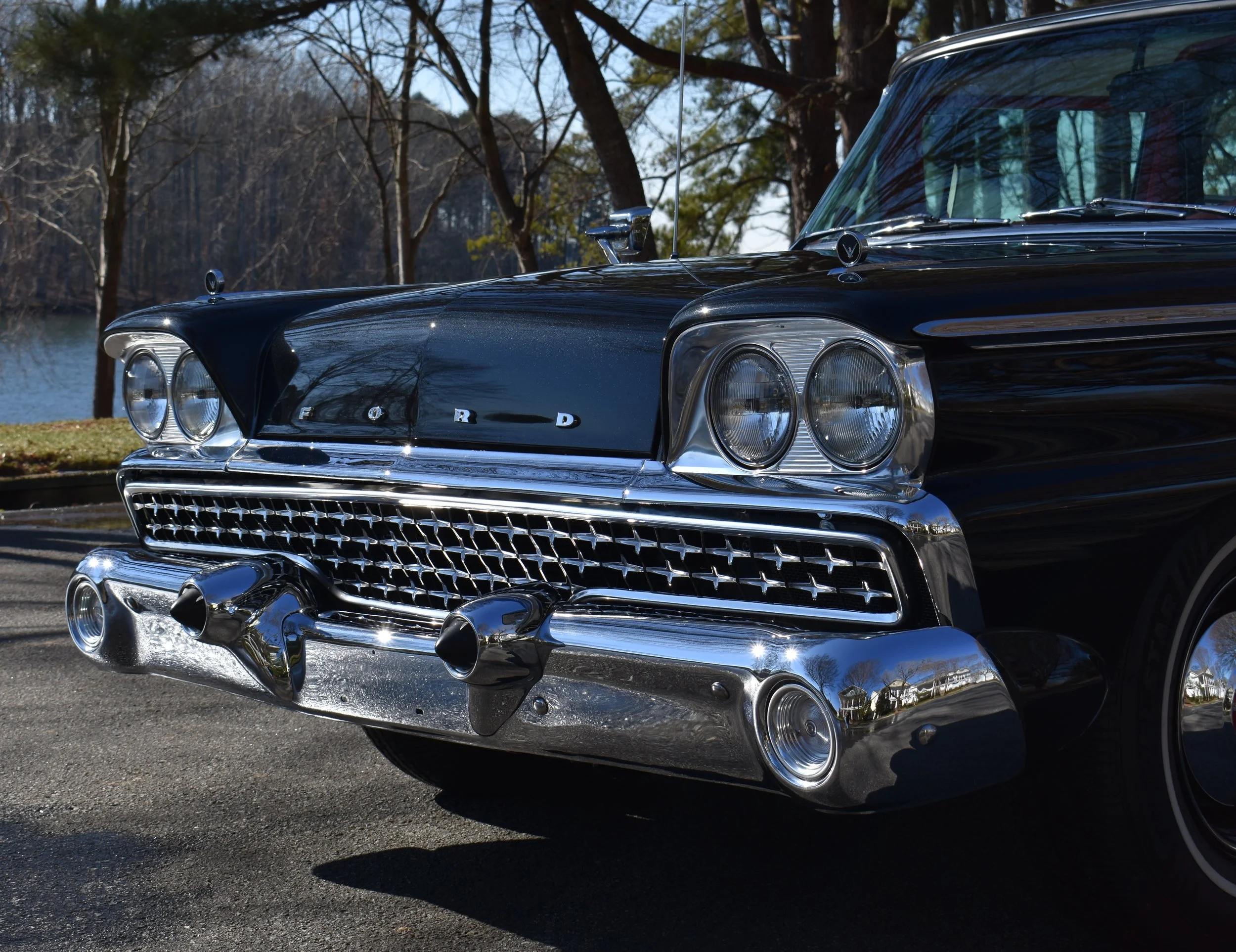 Close-up of a classic black Ford car with chrome details parked outdoors, with trees and a lake in the background.