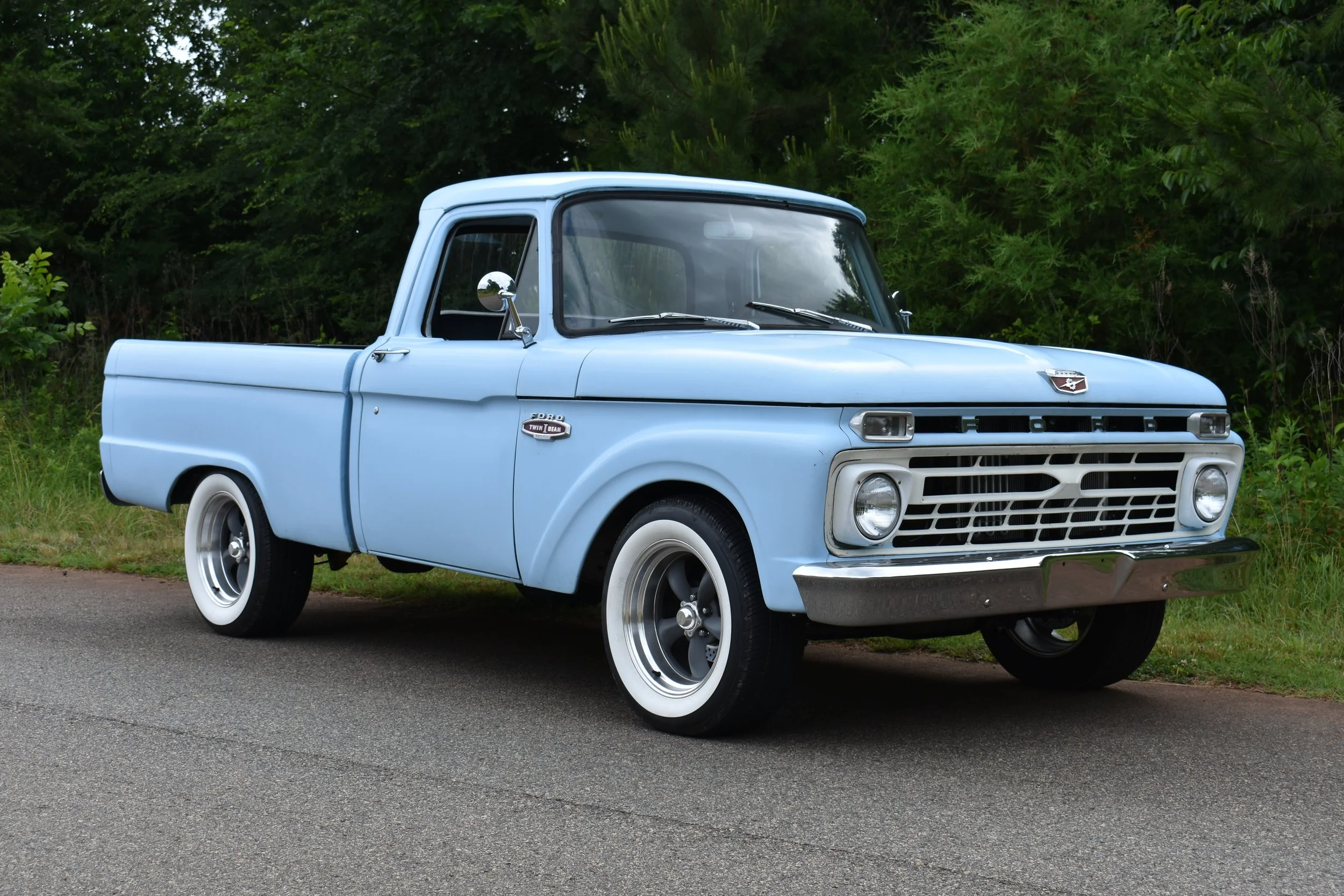 Light blue vintage pickup truck parked on a road with green bushes and trees in the background.