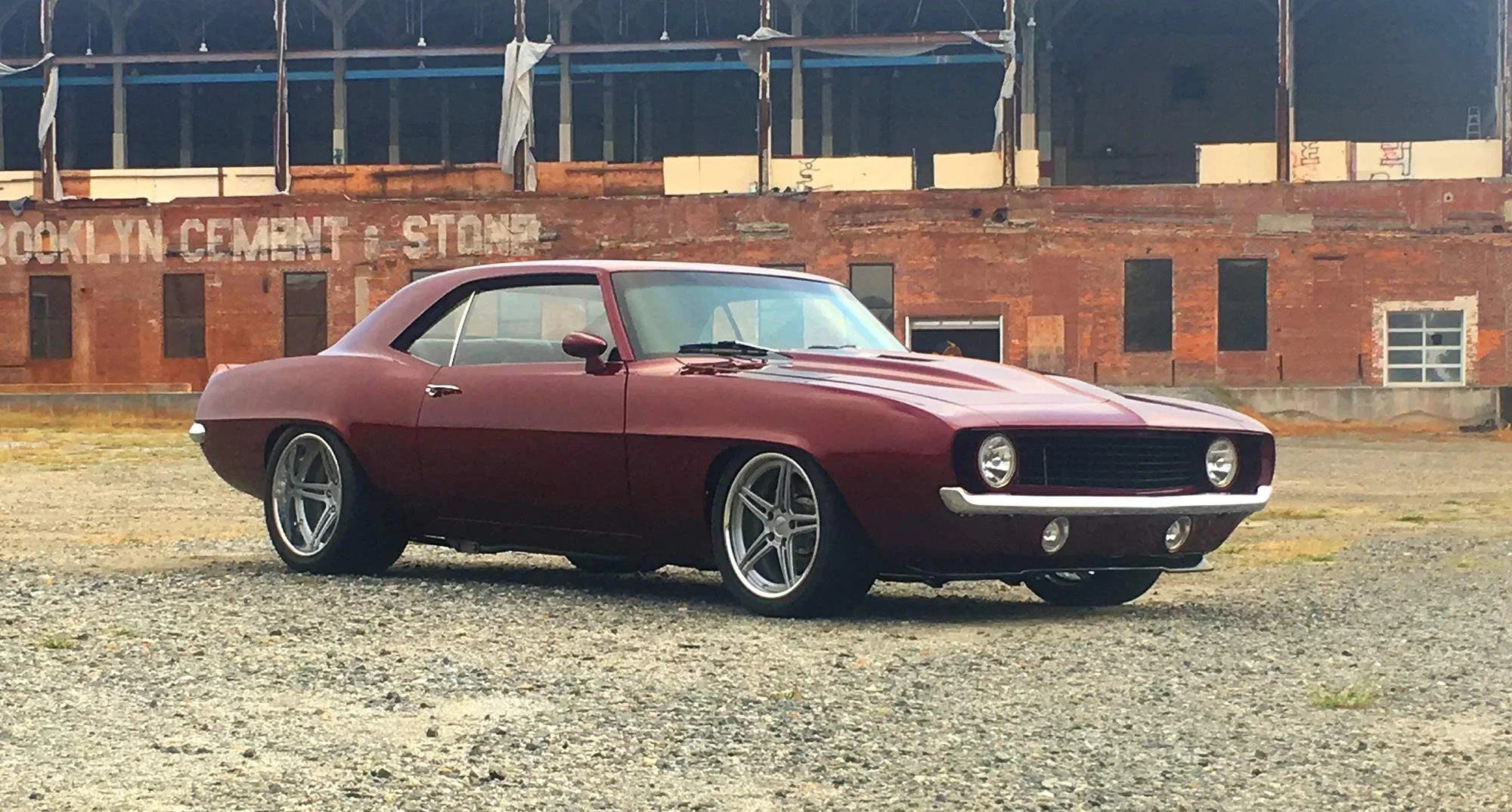 A vintage red sports car with a sleek, curved body parked on a gravel lot in front of an old brick building with partially covered windows and a sign that reads 'Brooklyn Cement & Stone.'
