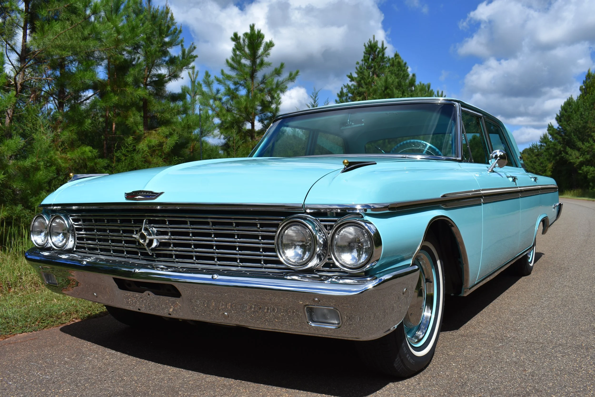 A vintage light blue car parked on a paved road, surrounded by green pine trees under a partly cloudy sky.