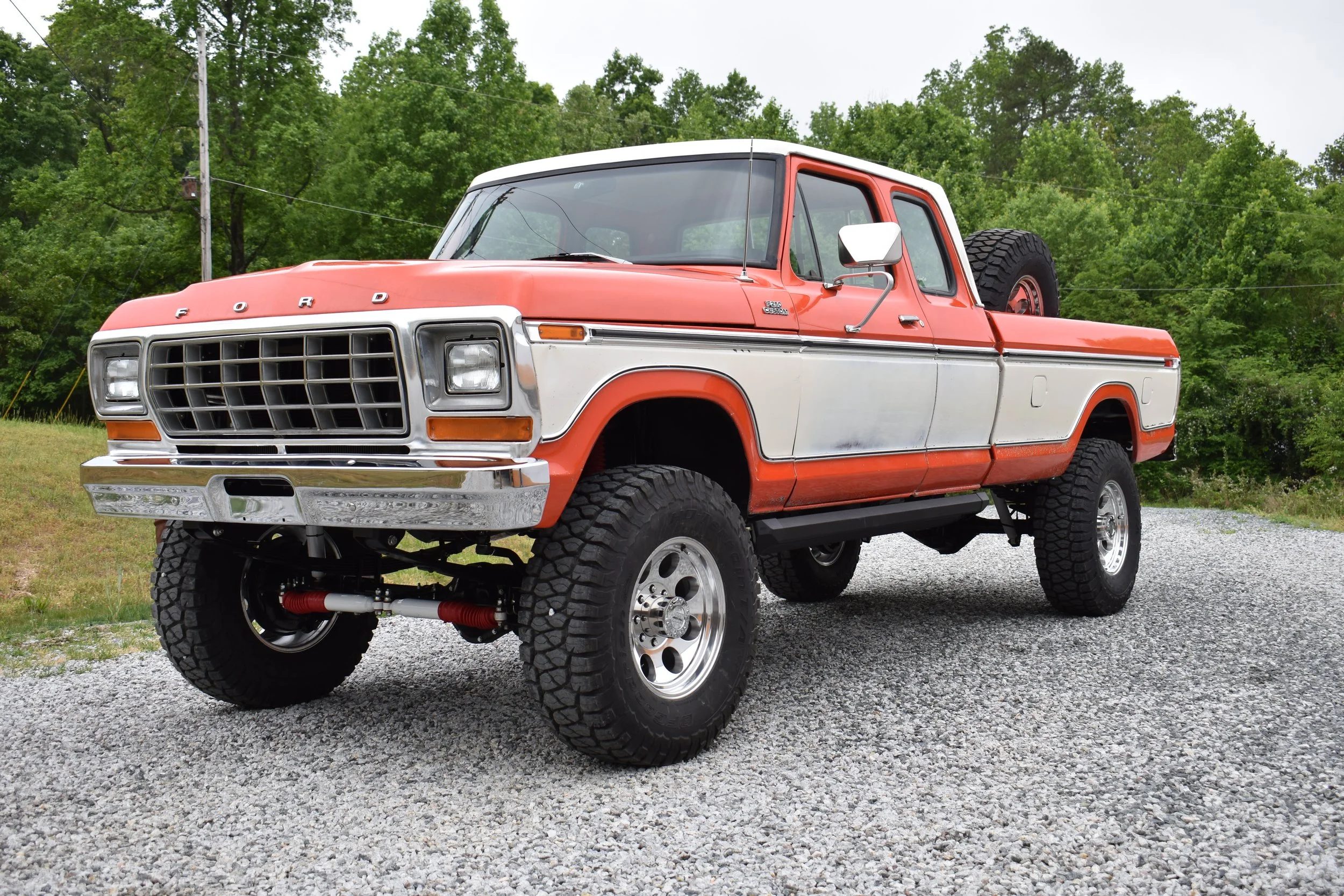 A vintage orange and white Ford pickup truck with large off-road tires parked on gravel in front of green trees.