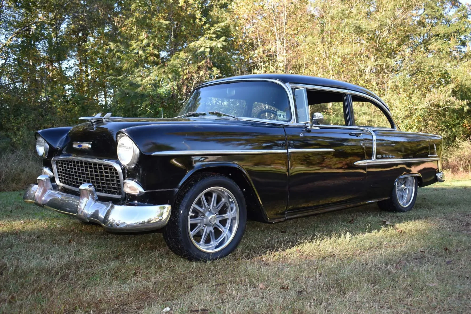 A vintage black Chevrolet Bel Air car parked on grass with a backdrop of trees and foliage.