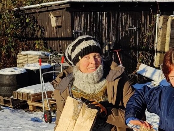 En kvinde med en strikket hue og tørklæde, der holder et stykke træ, udenfor i vinterlandskab.