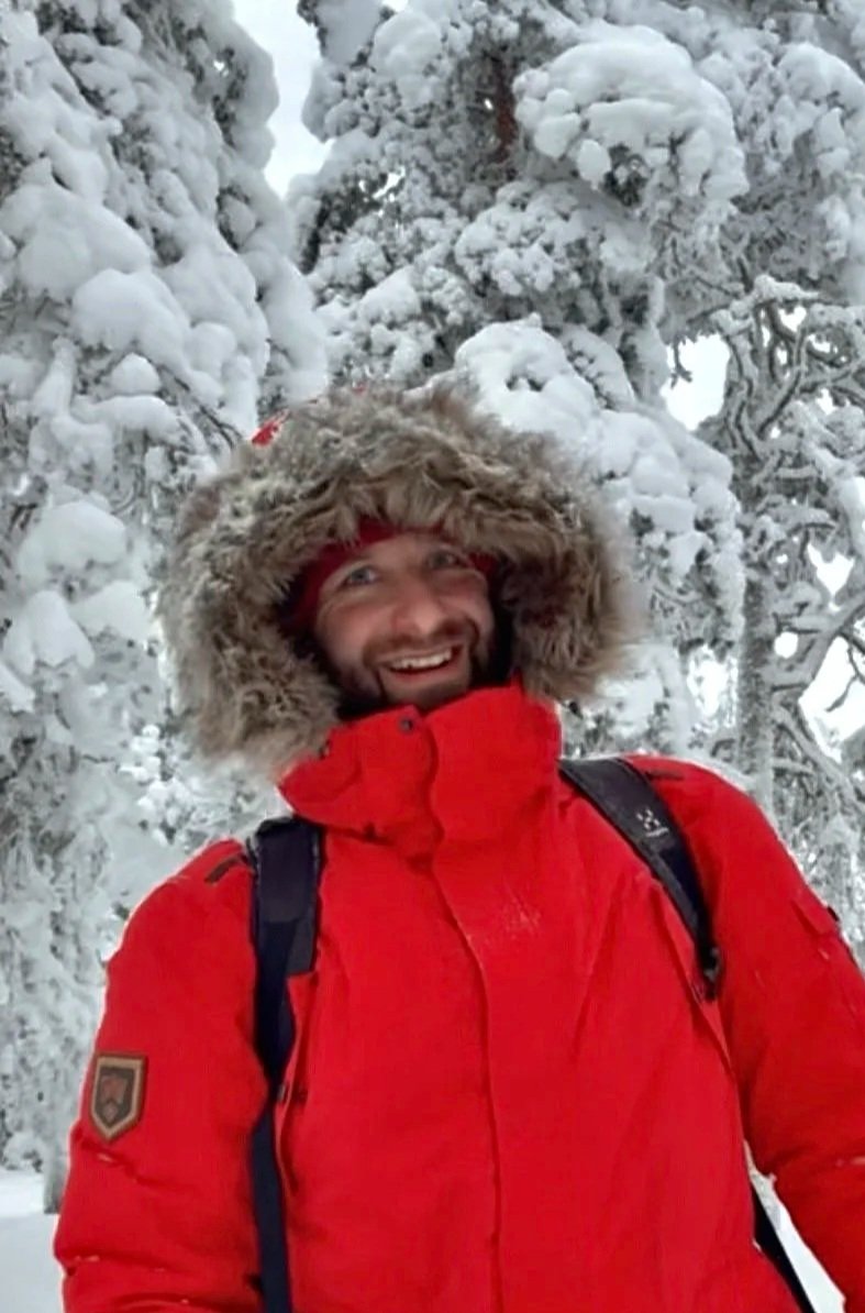 En mand i rød jakke og med pelsfrakkehat står foran sneklædte træer i snevejr og smiler.