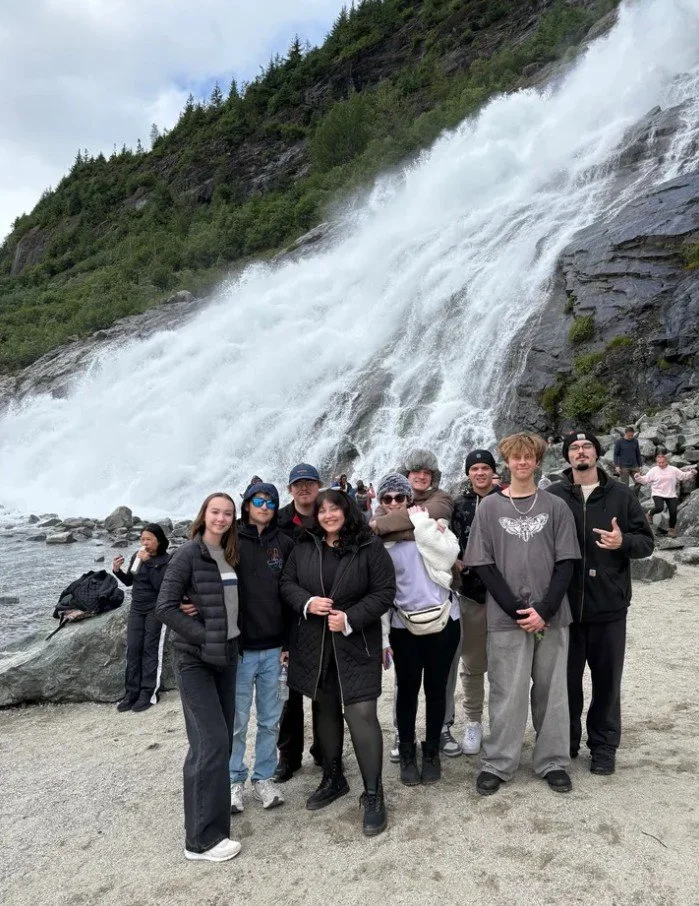 Group of people standing near a river with a large waterfall in the background.