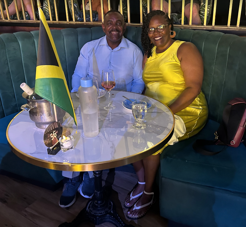 A man and woman sitting at a table in a restaurant, smiling at the camera. The man is wearing a light blue shirt and the woman is in a yellow dress. There are drinks on the table, a small flag, and a purse placed on the seat.