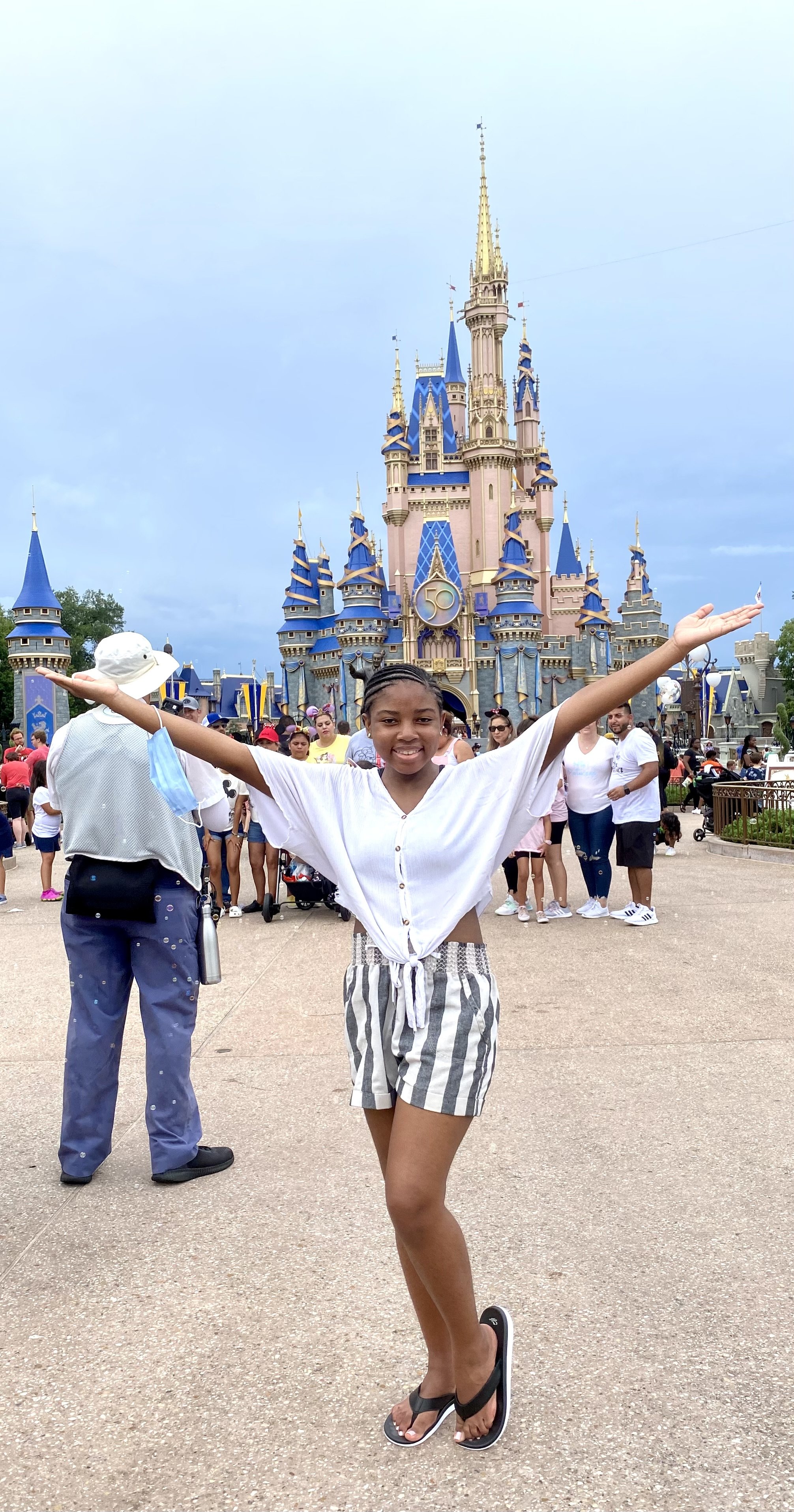 A young girl smiling with arms outstretched in front of Cinderella's Castle at Disney World, with a crowd of people behind her.