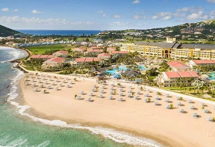 Aerial view of a beach resort with sandy beach, lounge chairs under umbrellas, a hotel complex with pools, and a coastal landscape with hills and ocean in the background.