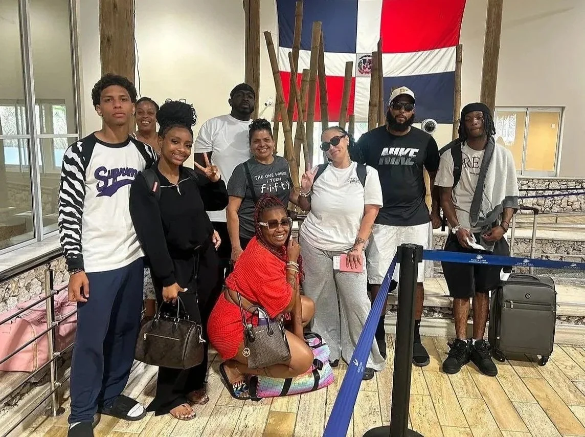 Group of people posing inside an airport, with a large flag of the Dominican Republic hanging behind them, and a blue rope barrier in front.